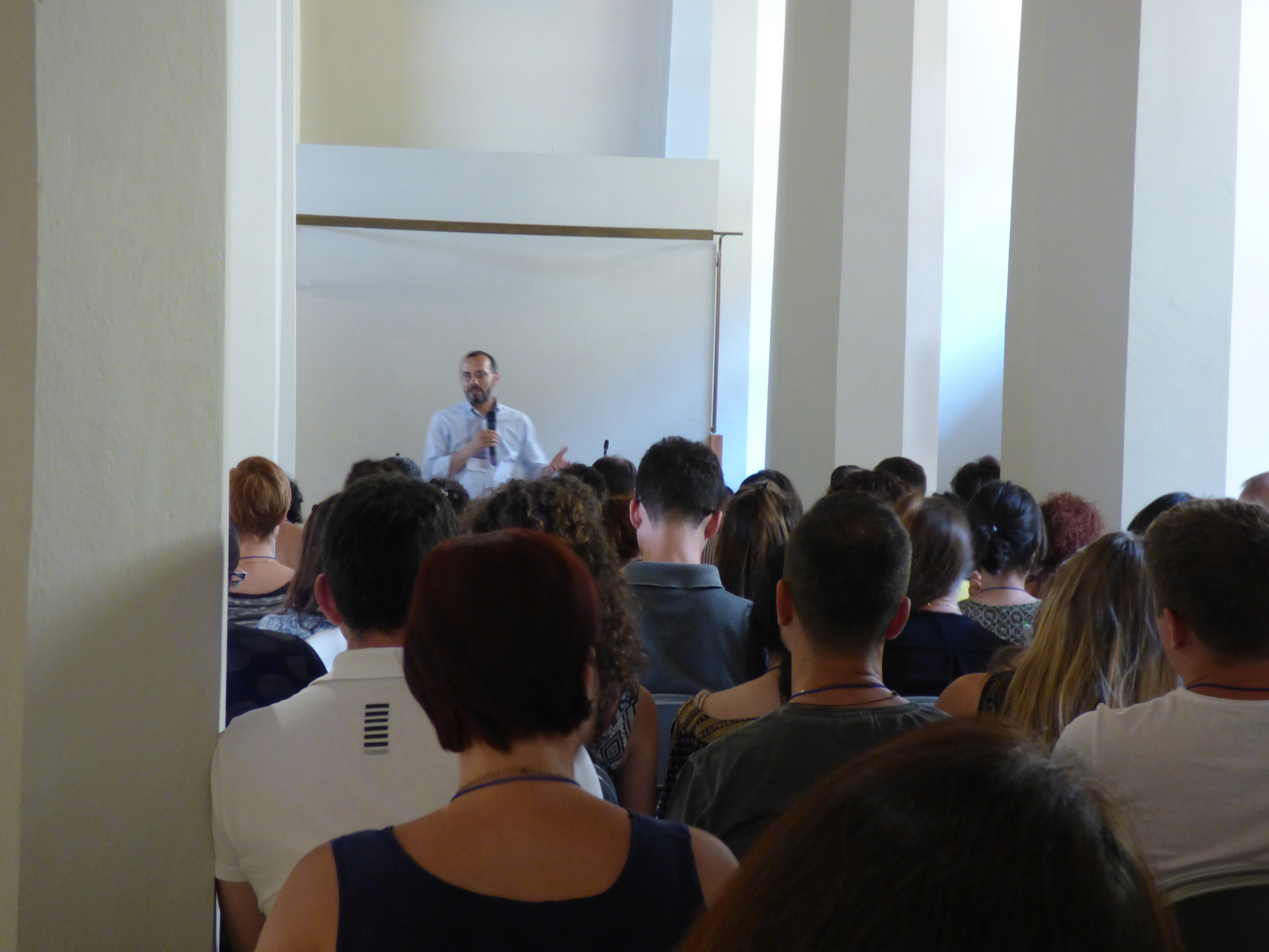 Un relatore parla in fondo all'aula, ripreso da dietro il pubblico seduto.