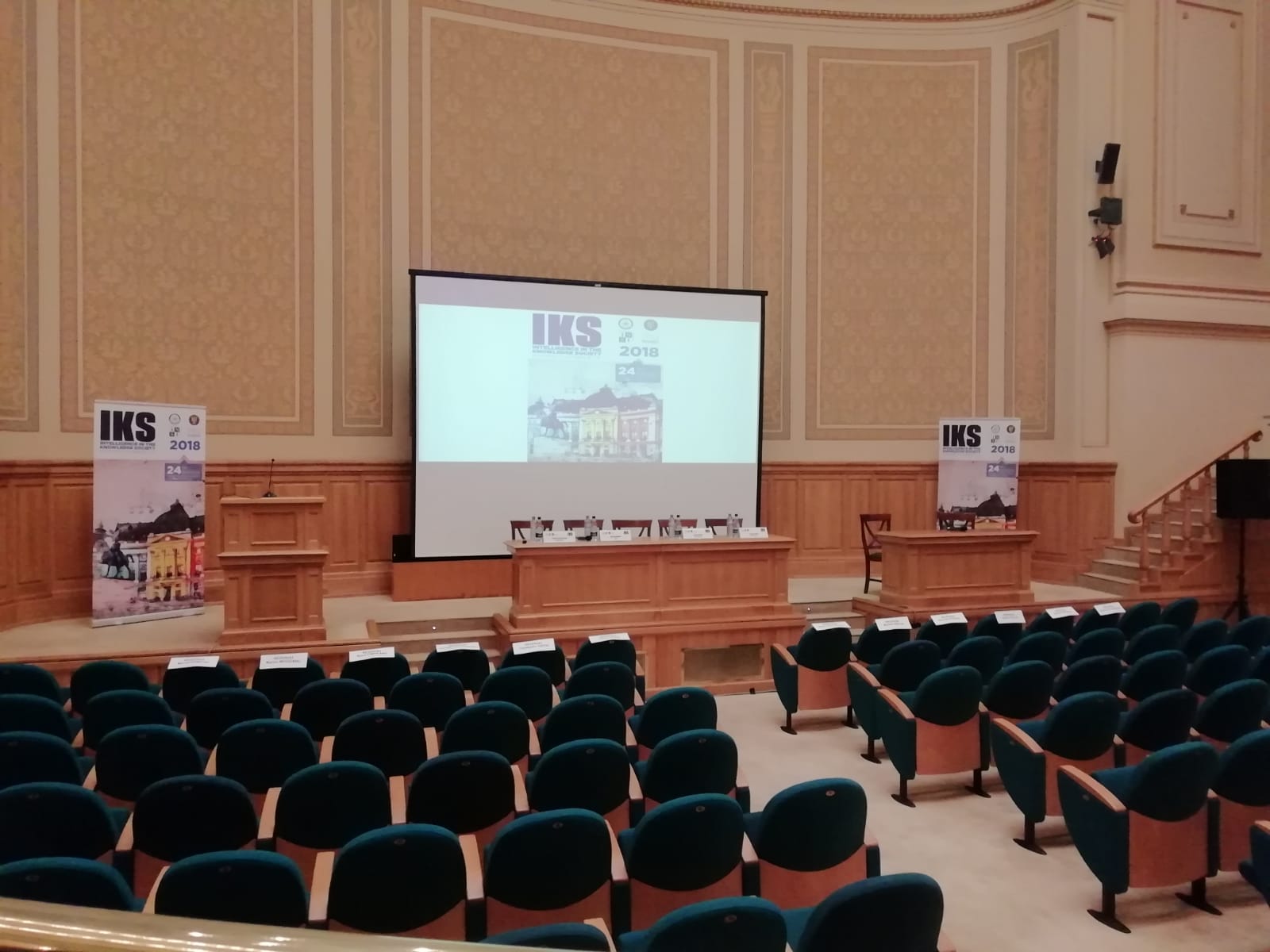 Un'aula magna vuota pronta per la conferenza IKS: le poltrone verdi, lo schermo e i banner.