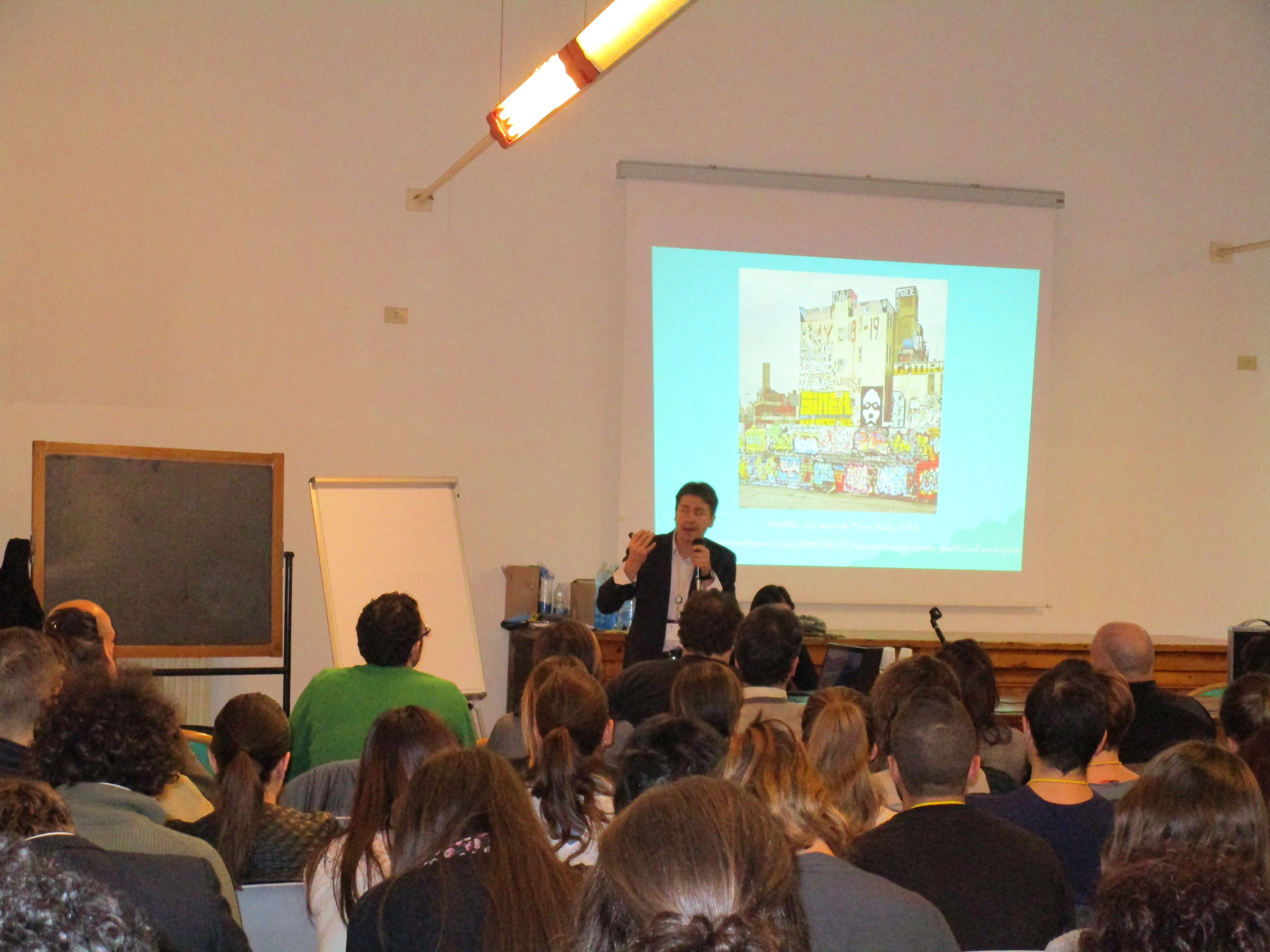 Un relatore davanti alla proiezione di un'illustrazione, in un'aula con il pubblico seduto.
