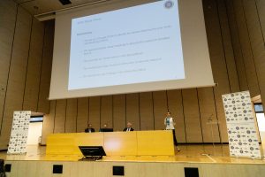 Un'aula magna con il tavolo dei relatori e la proiezione di una diapositiva.