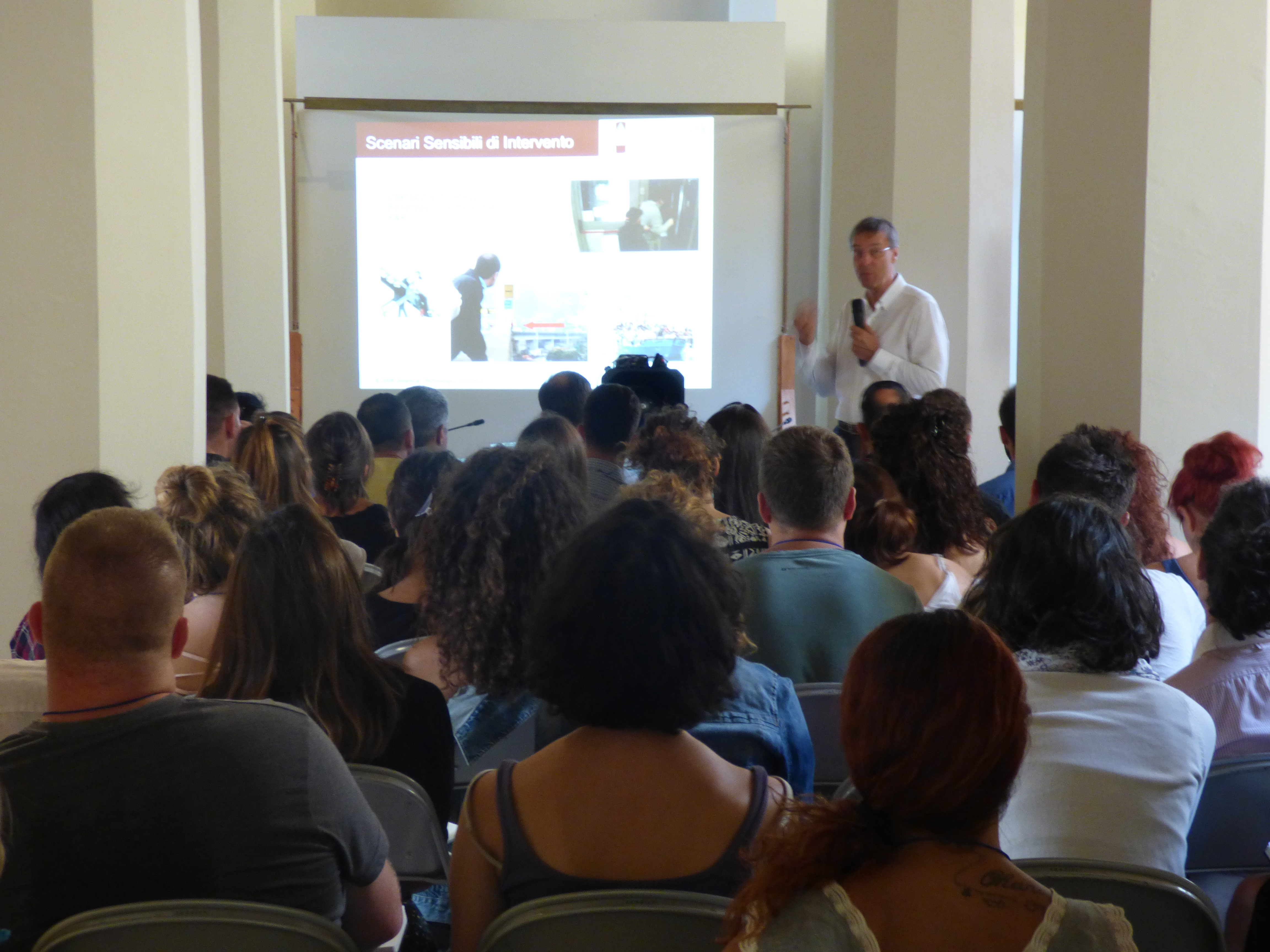 Un relatore accanto alla proiezione di una diapositiva sugli scenari di intervento, davanti a un'aula piena.