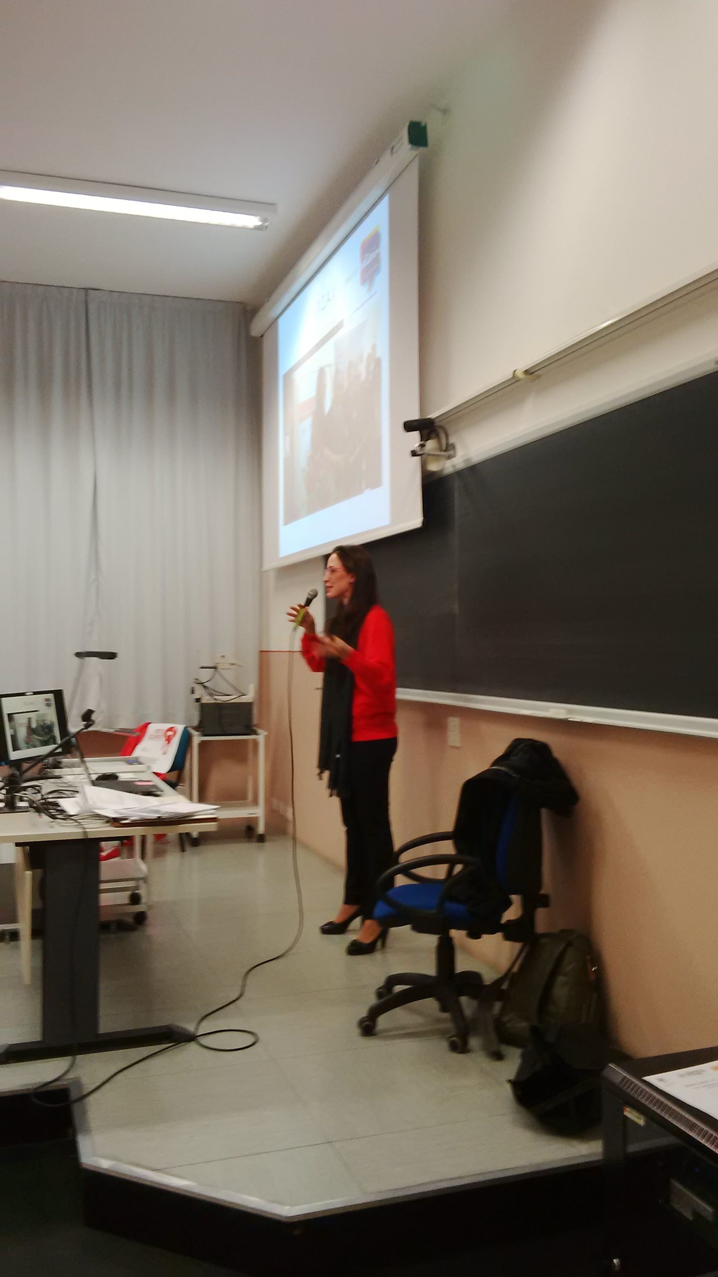 Una relatrice al microfono accanto alla lavagna e alla proiezione di una fotografia.