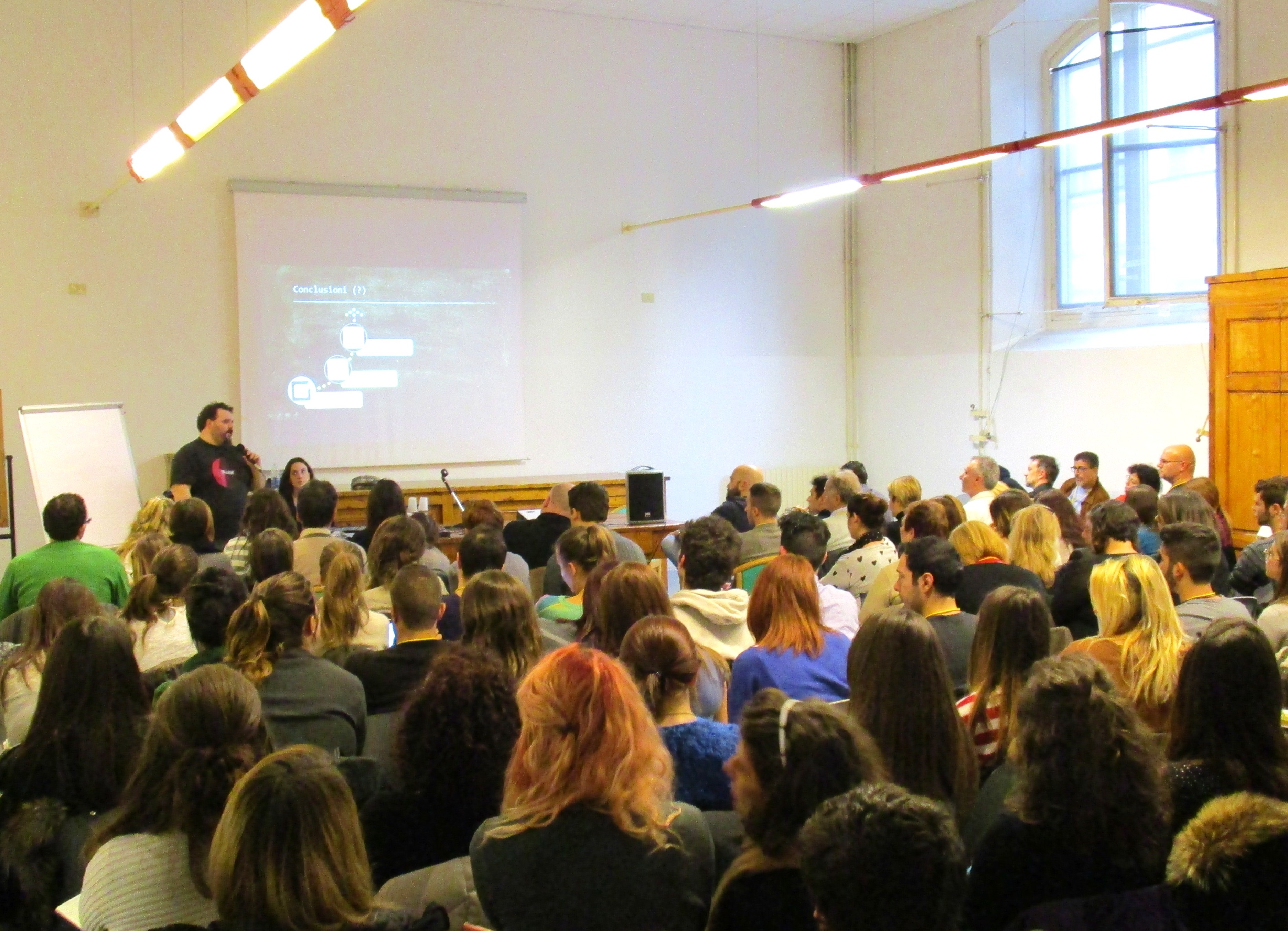 L'aula piena vista da dietro, con due relatori in fondo accanto allo schermo.