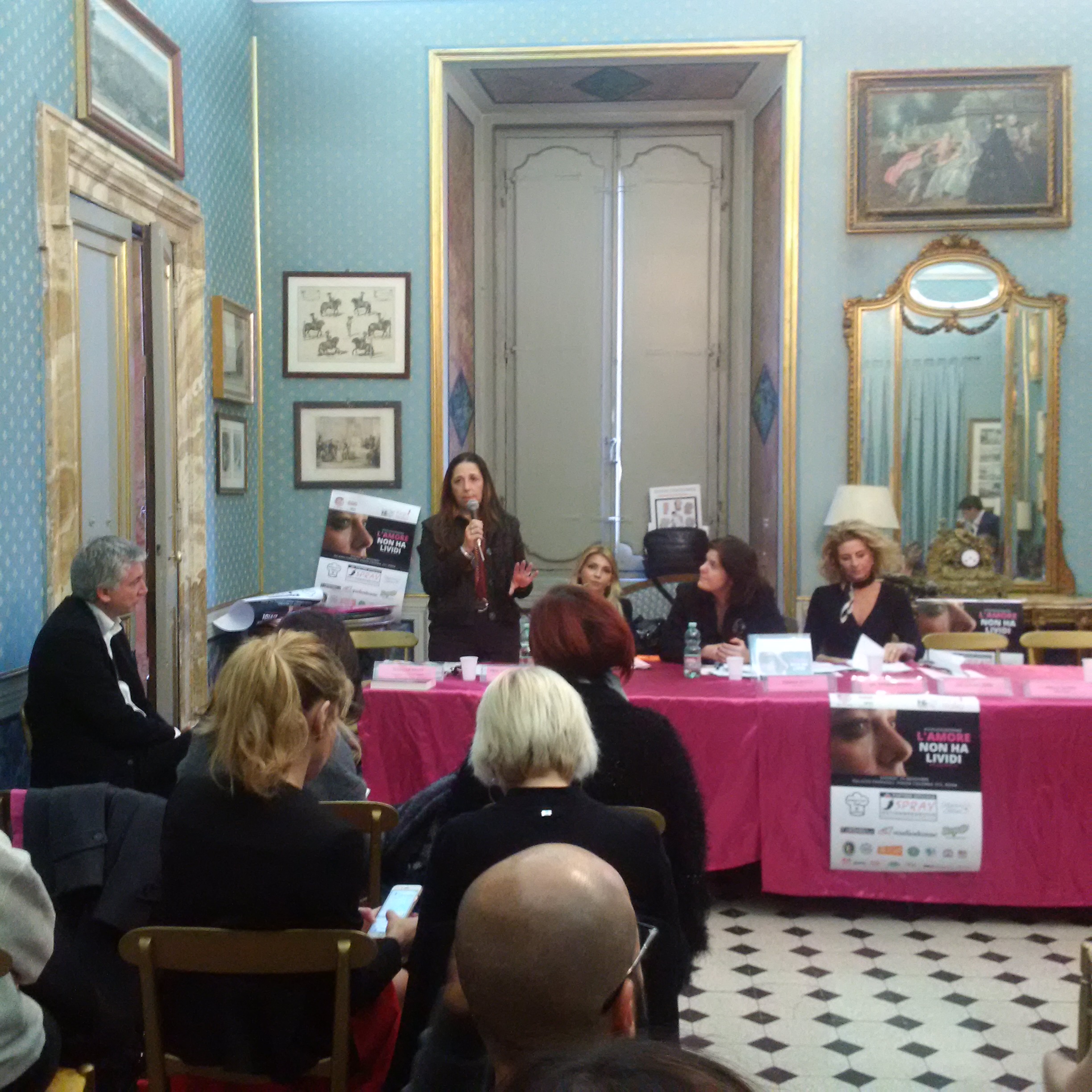 Una relatrice in piedi parla al tavolo delle relatrici, in una sala storica dal pavimento a scacchi.