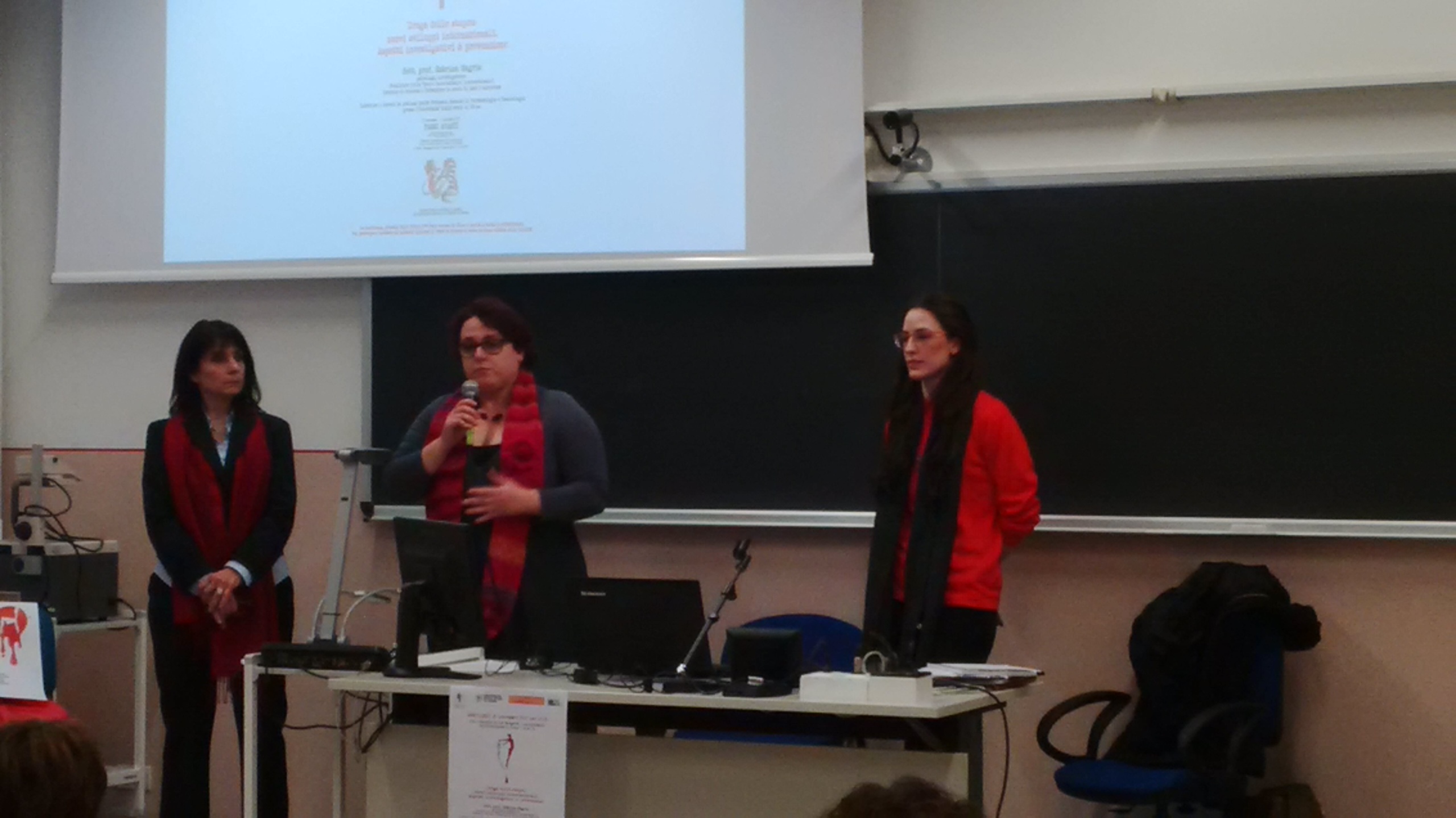 Tre relatrici in piedi davanti alla lavagna e alla proiezione della locandina.