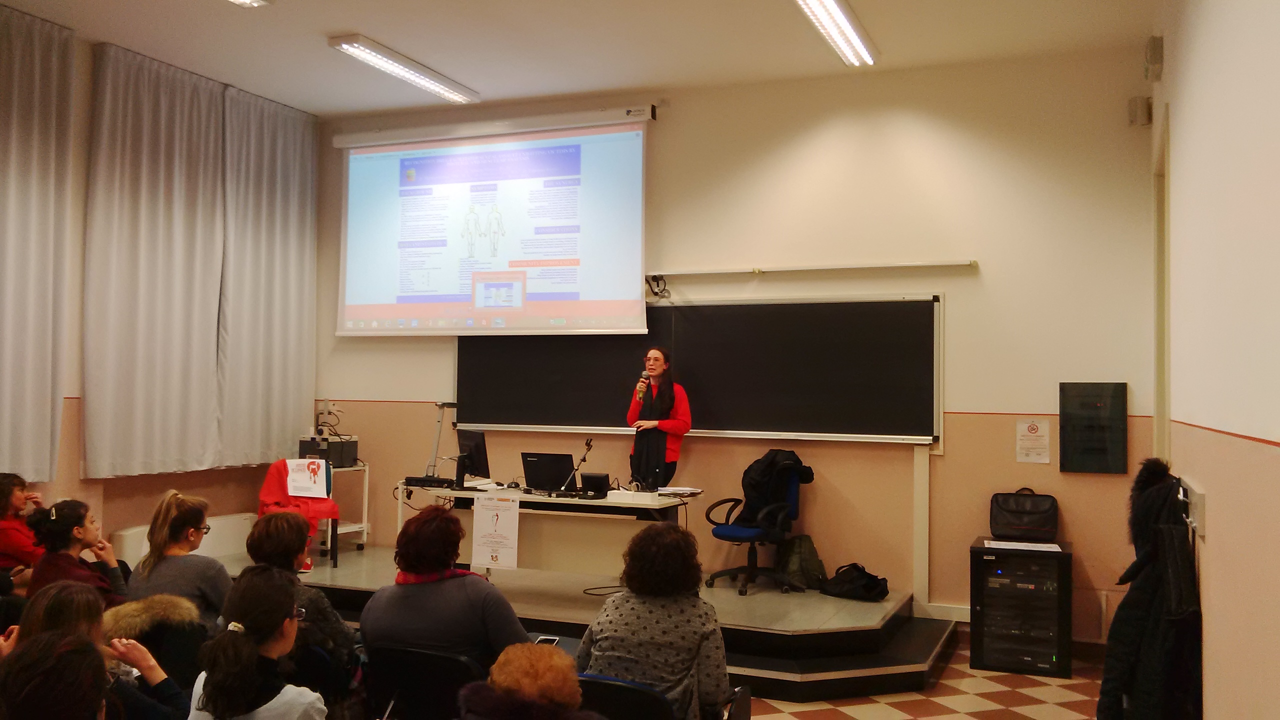 Una relatrice sulla pedana accanto alla proiezione, davanti al pubblico seduto.