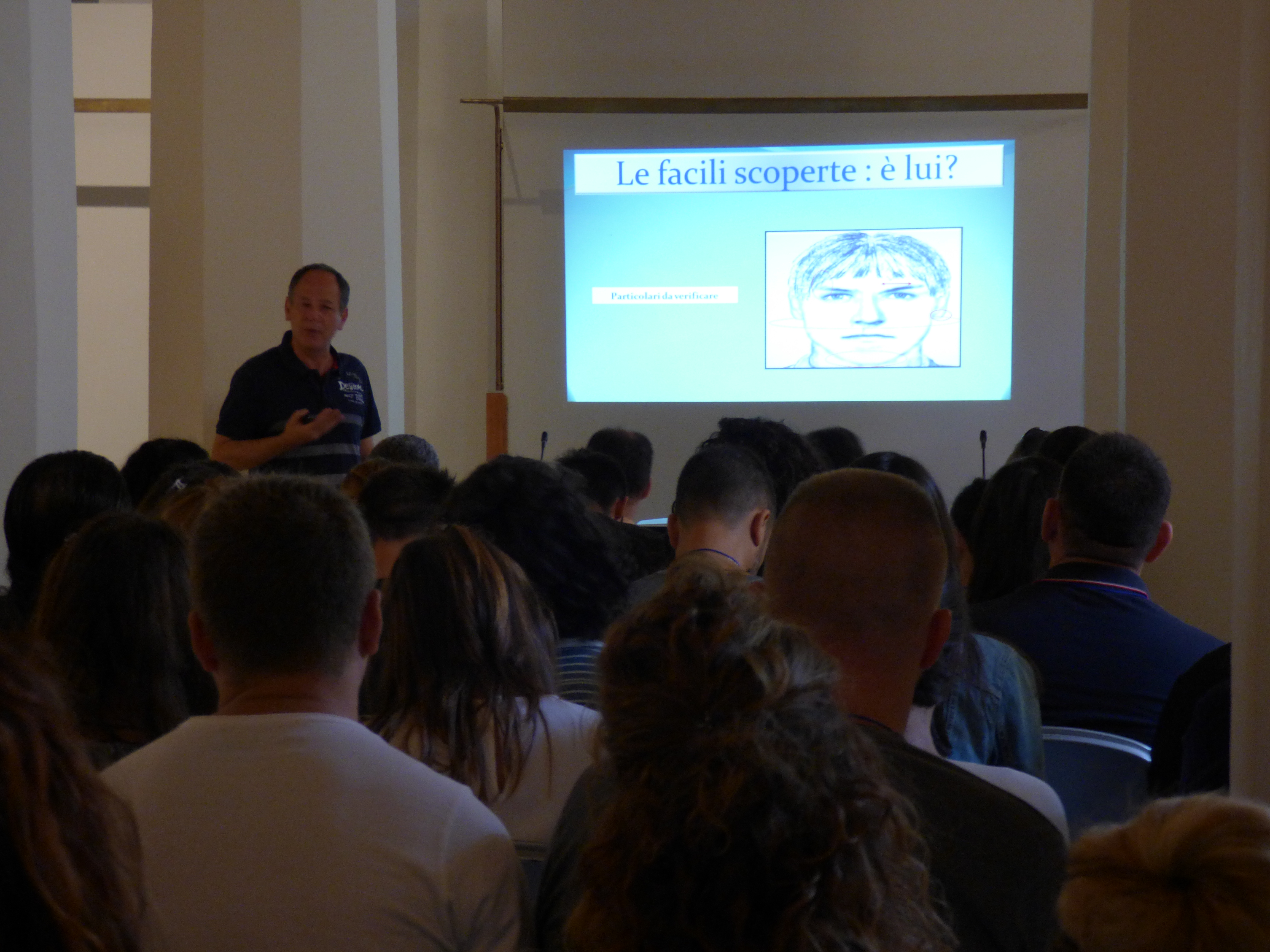 Un relatore accanto alla proiezione intitolata Le facili scoperte: è lui?, davanti al pubblico seduto in un'aula chiara.