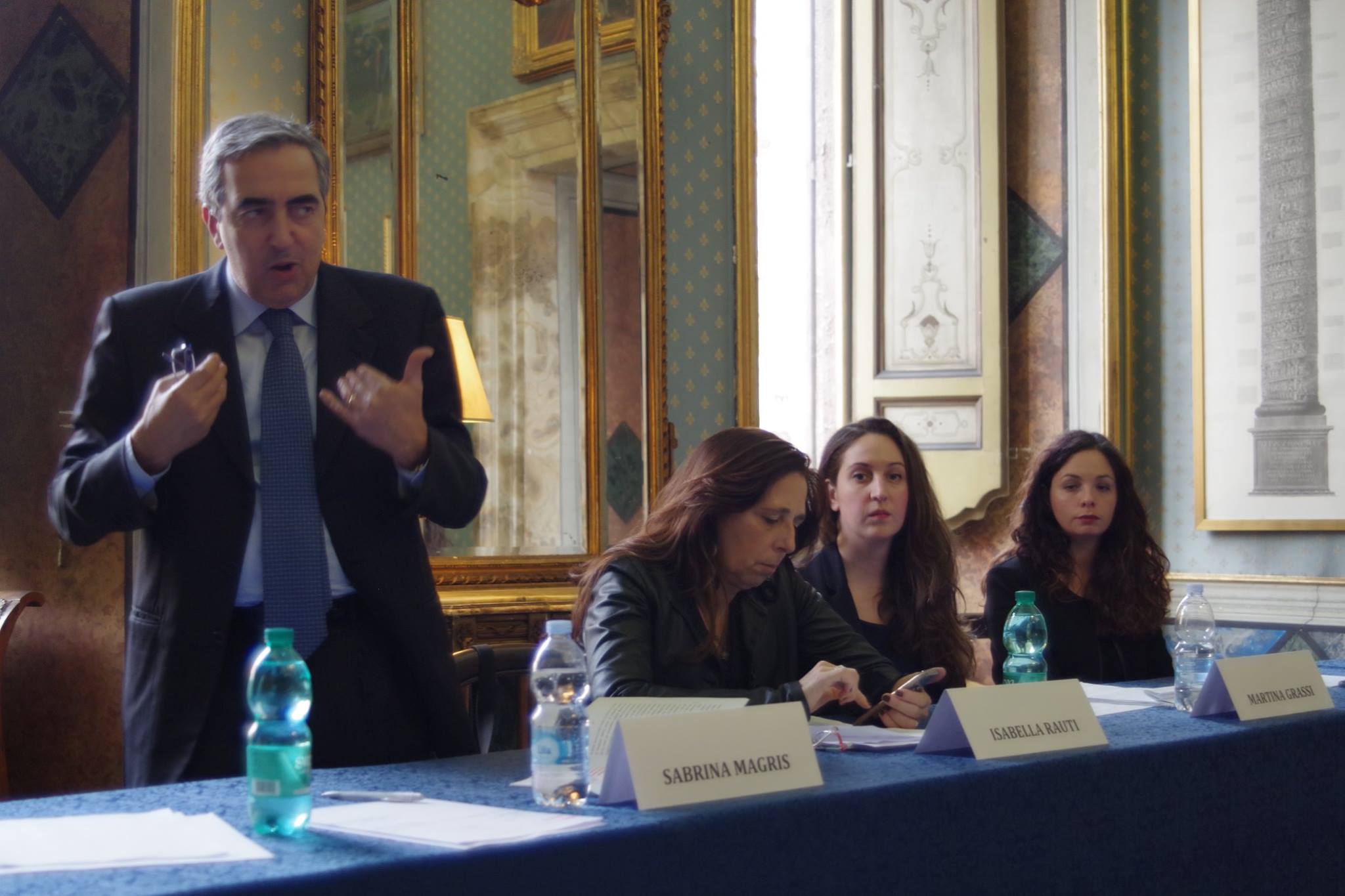 Un relatore in piedi parla al tavolo delle relatrici, in una sala affrescata.