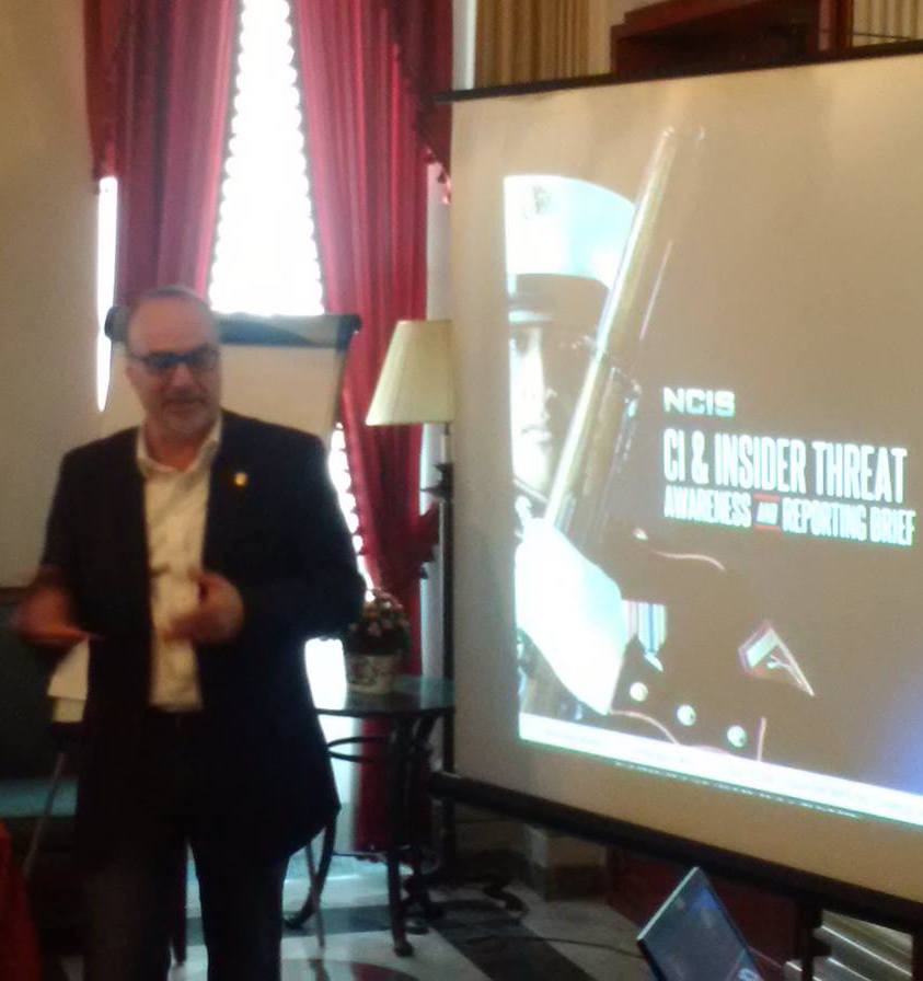 Un relatore accanto alla proiezione NCIS, CI and Insider Threat Awareness and Reporting Brief, in una sala dalle tende rosse.