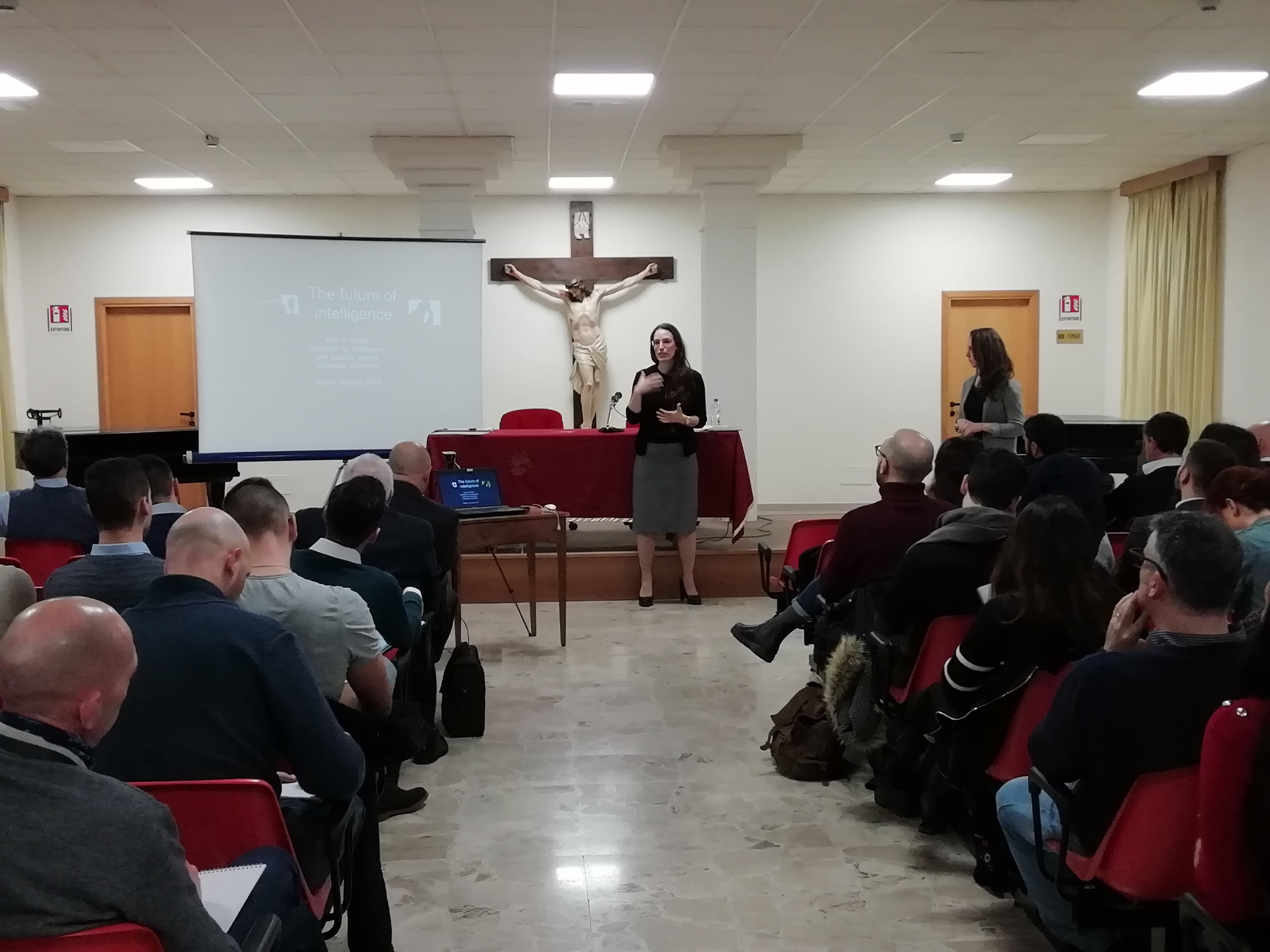 Una relatrice in piedi sulla pedana davanti al pubblico seduto, sotto un crocifisso.