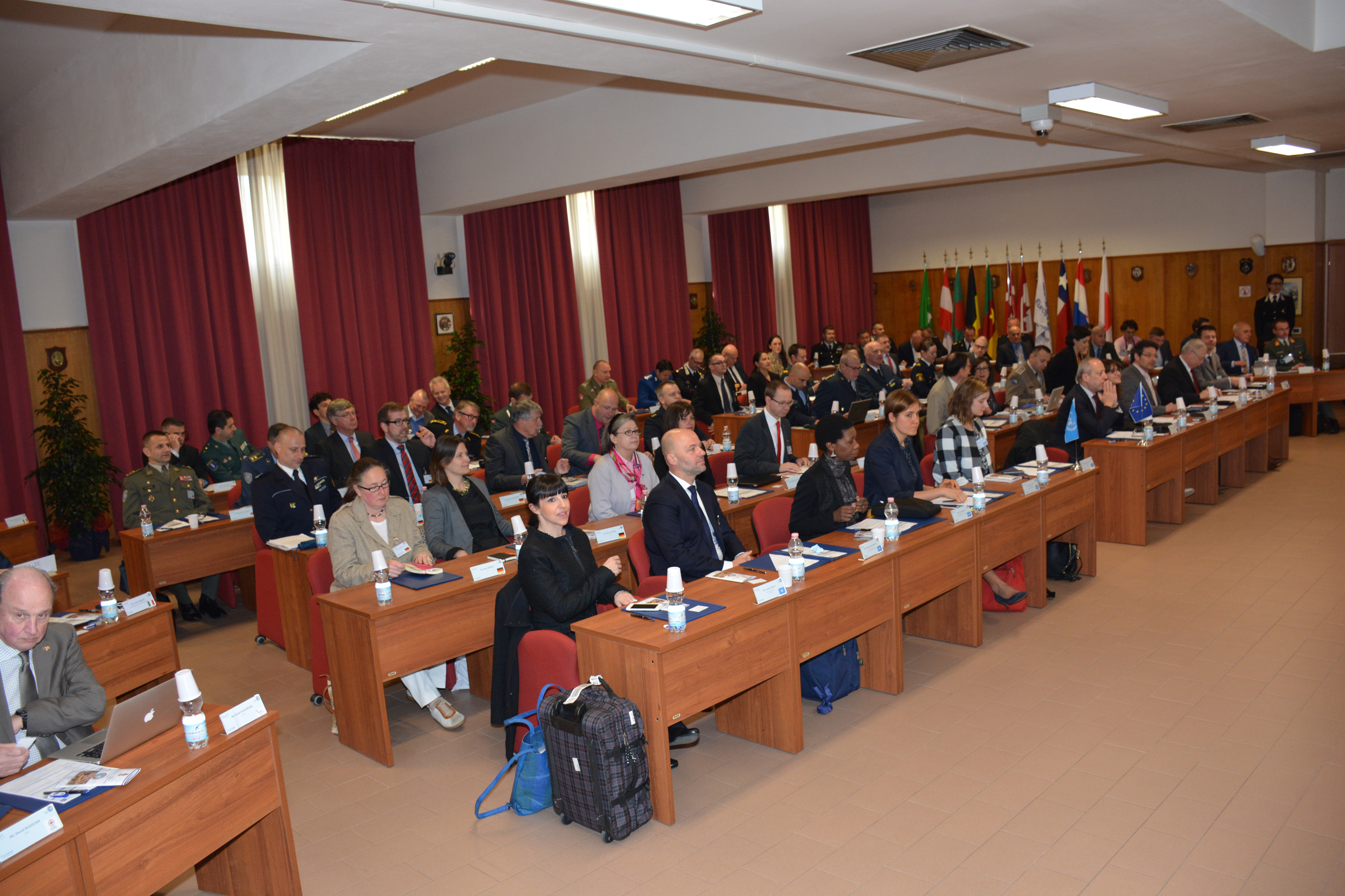 Un'aula con i banchi pieni di partecipanti, alcuni in uniforme, e le bandiere in fondo alla sala.