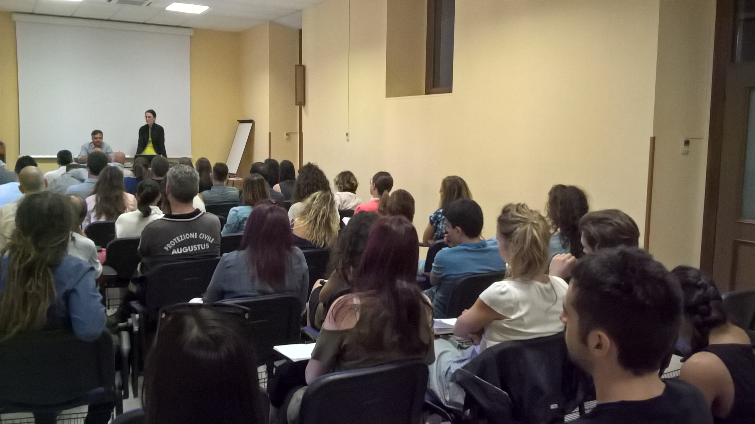 Un'aula piena vista da dietro, con due relatori in fondo davanti allo schermo.