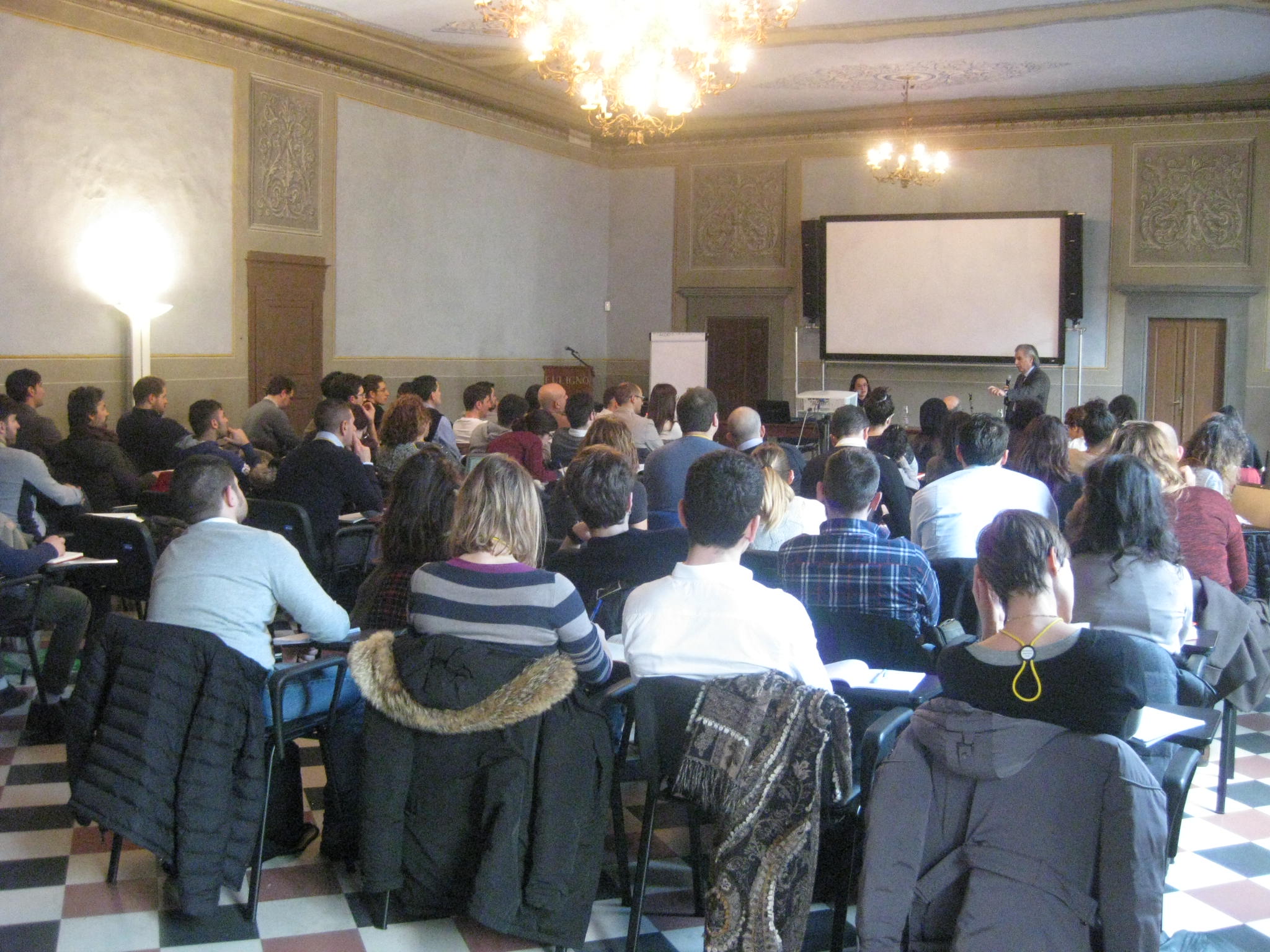 Il pubblico seduto in una sala storica con i lampadari, davanti allo schermo e a un relatore.