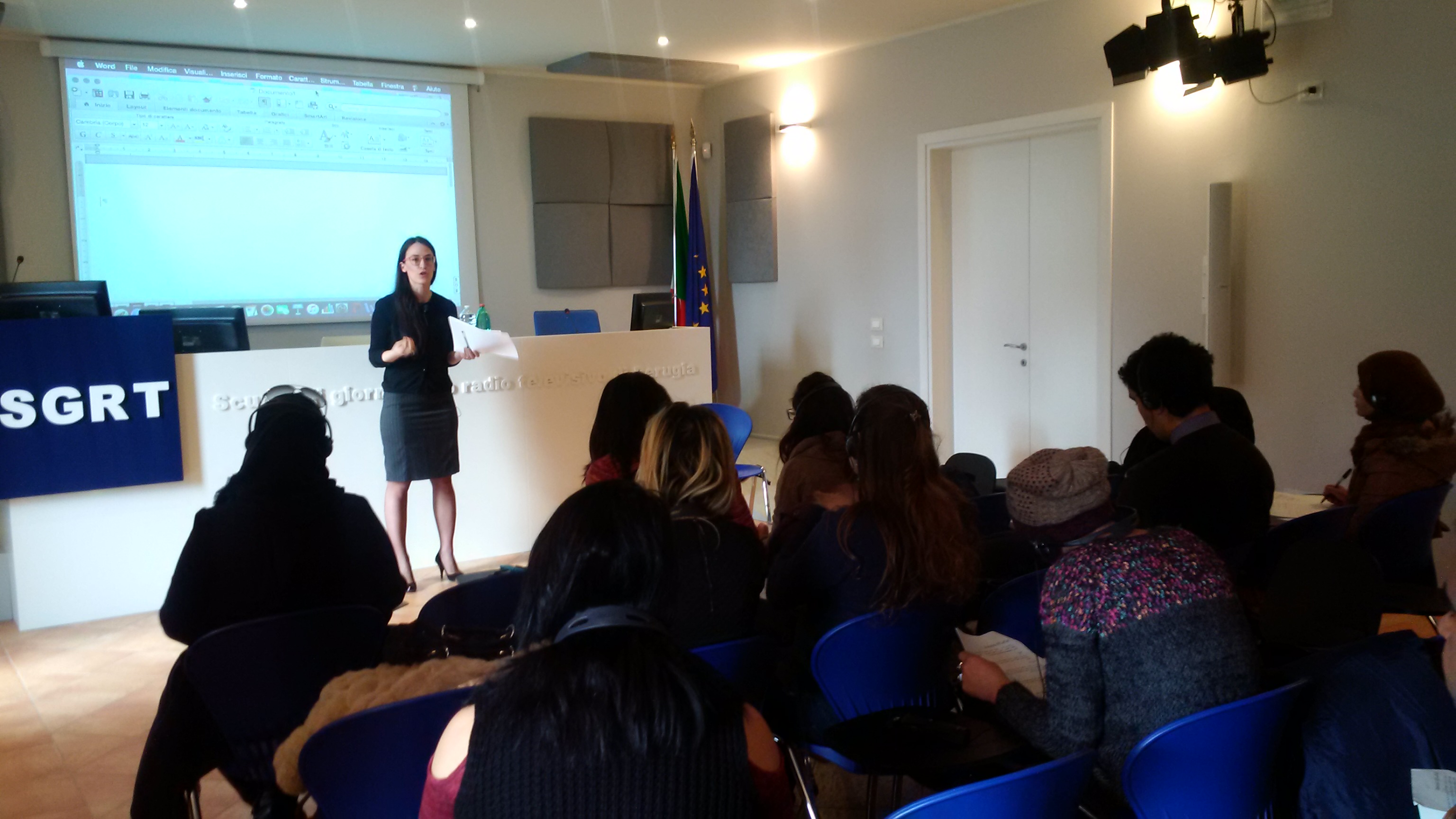 Una relatrice parla davanti a un'aula di partecipanti seduti, accanto alla bandiera italiana e a quella europea.