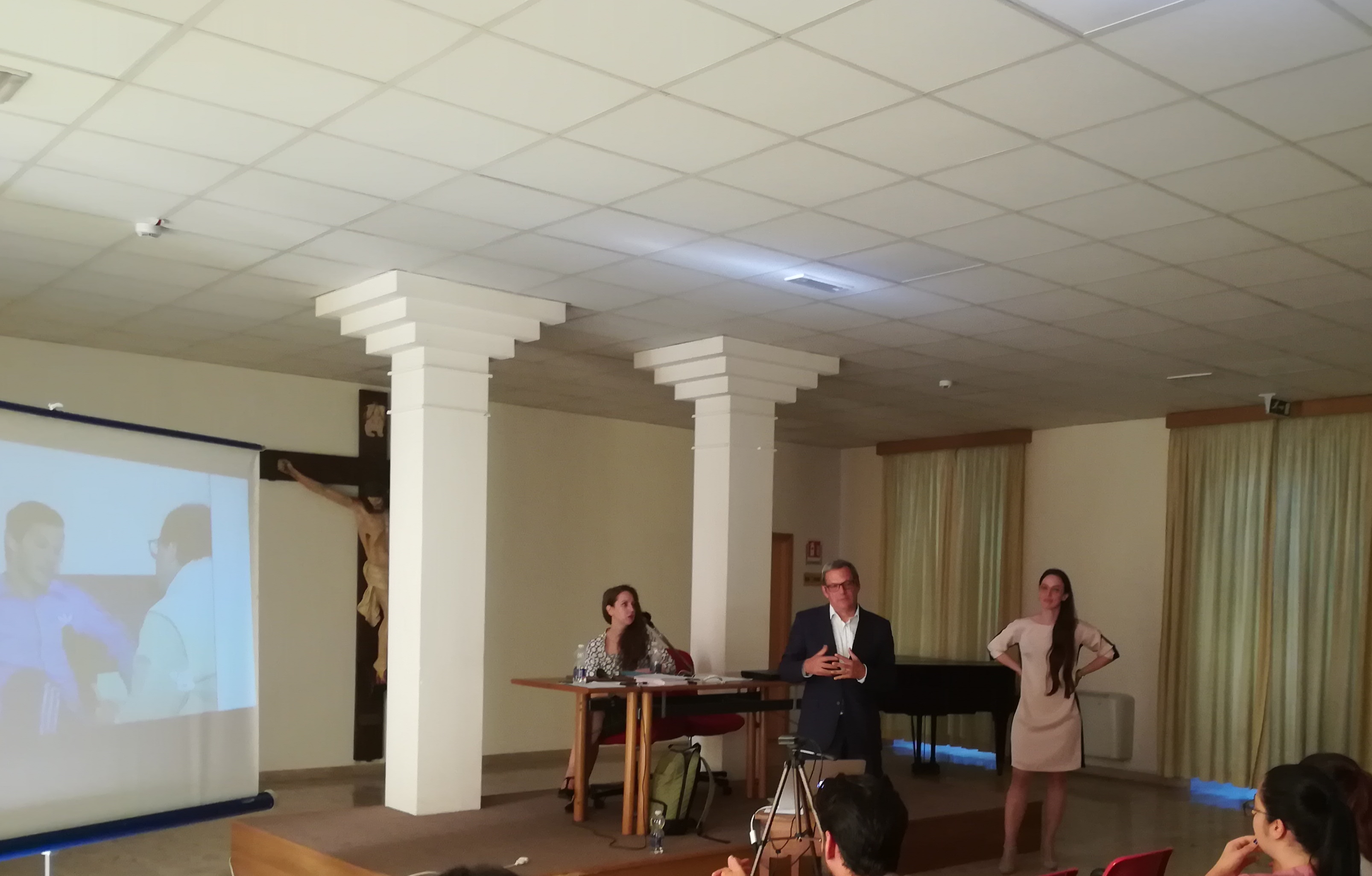 Due relatrici e un relatore davanti alla proiezione di un filmato, in una sala con le colonne.