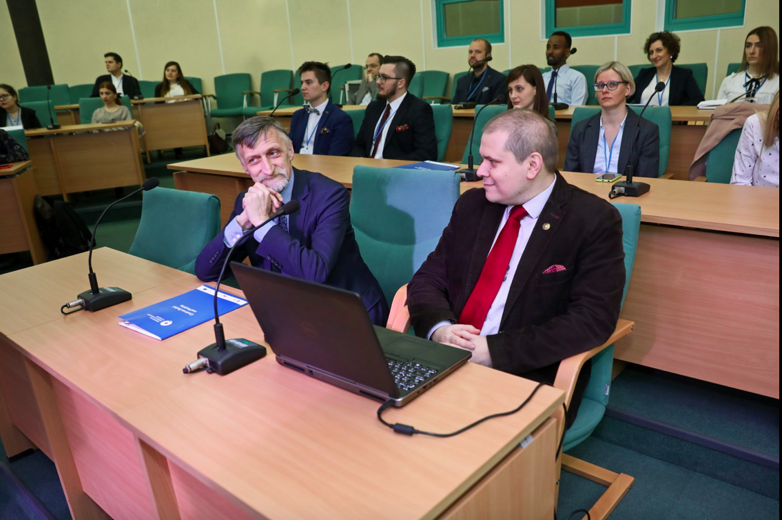 Due partecipanti seduti al banco con il portatile, in un'aula universitaria; dietro di loro altri partecipanti.