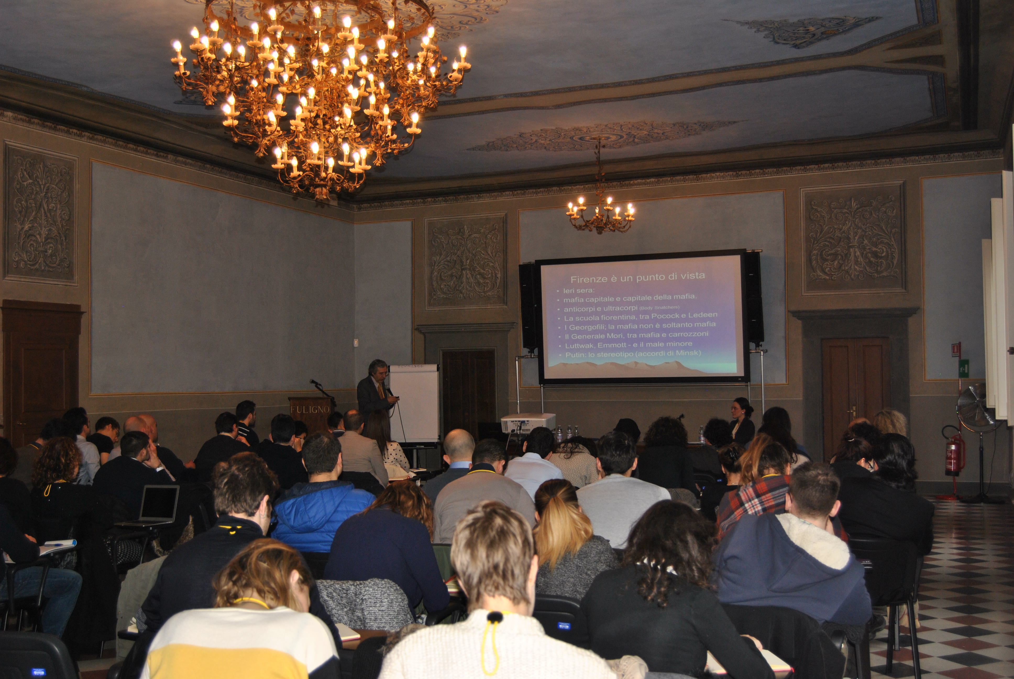 Una sala con i lampadari accesi, il pubblico seduto e la proiezione delle diapositive in fondo.