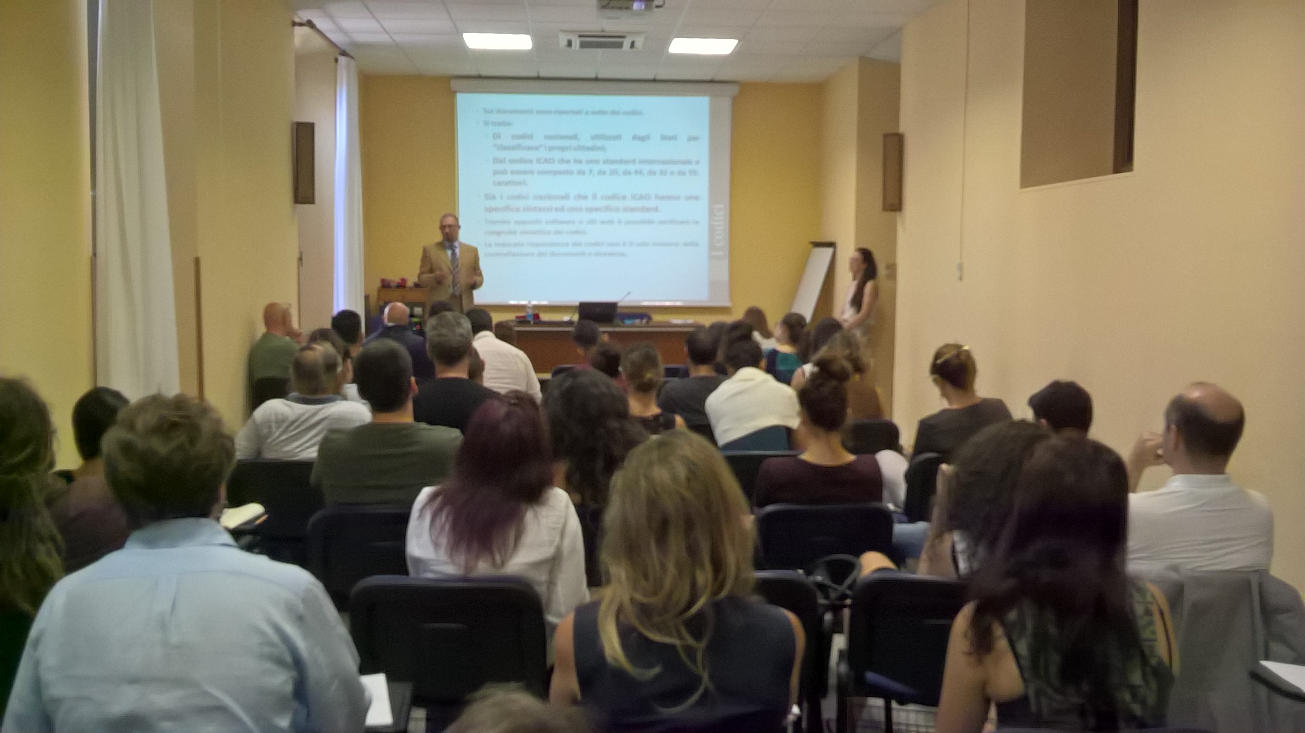 Un relatore parla in fondo all'aula accanto alla proiezione di un testo, con il pubblico seduto di spalle.