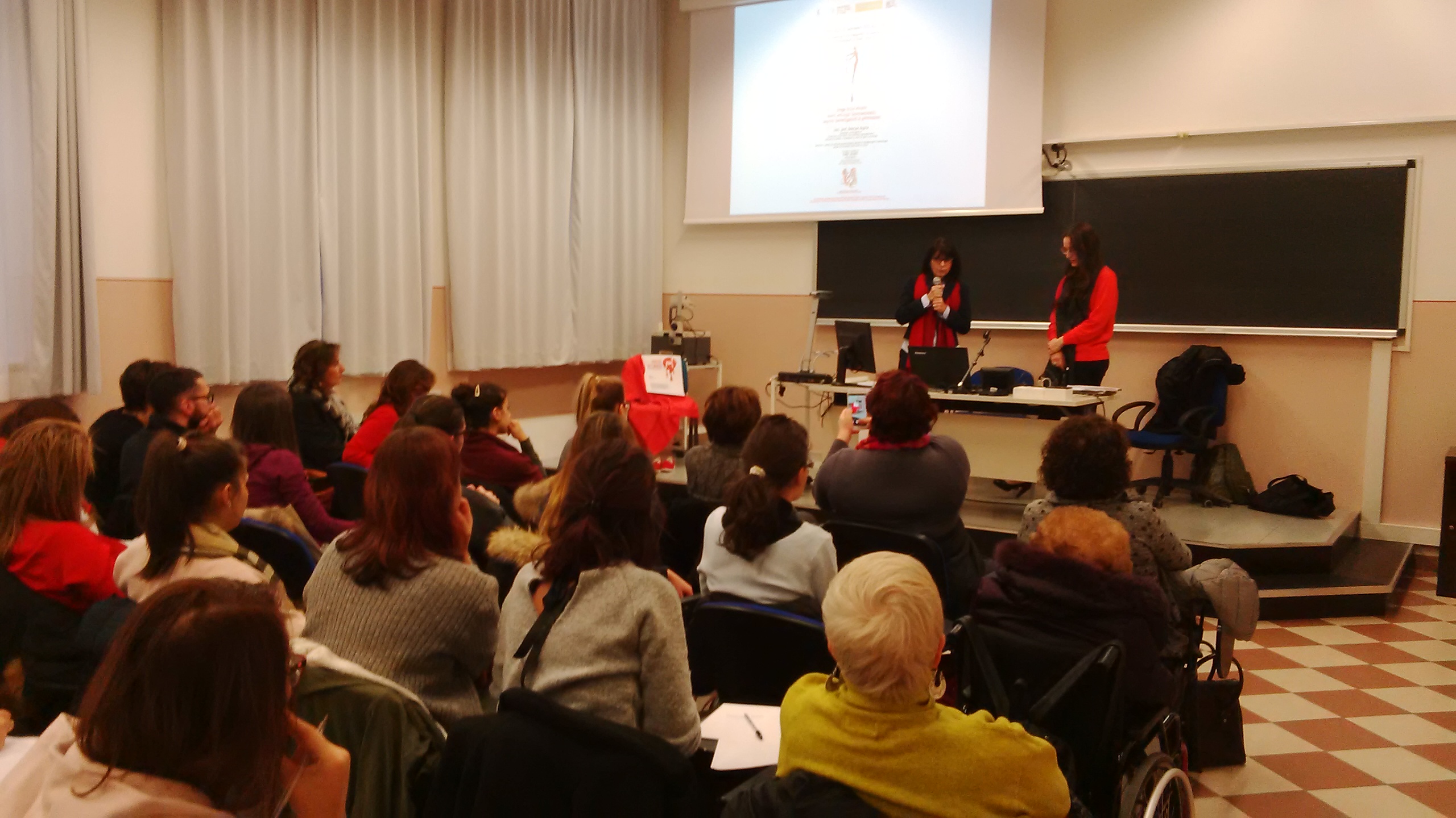 L'aula piena vista da dietro, con due relatrici sulla pedana accanto allo schermo.