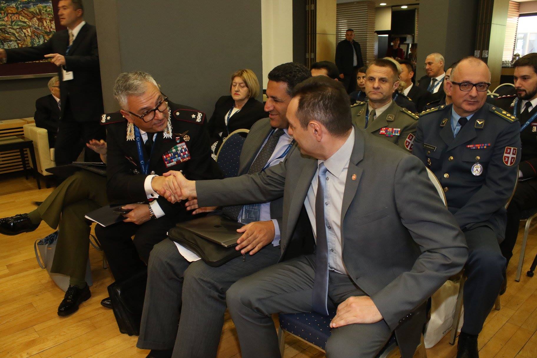 Due persone si stringono la mano fra le sedie della sala, in mezzo a ufficiali in uniforme.