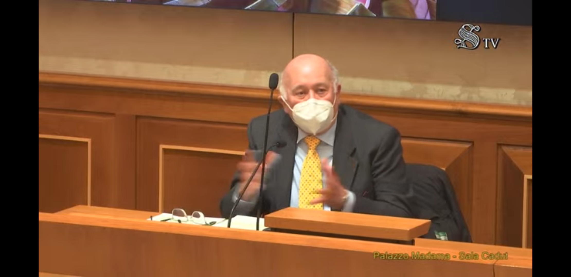 Un altro relatore con la mascherina parla al microfono, nella stessa sala.