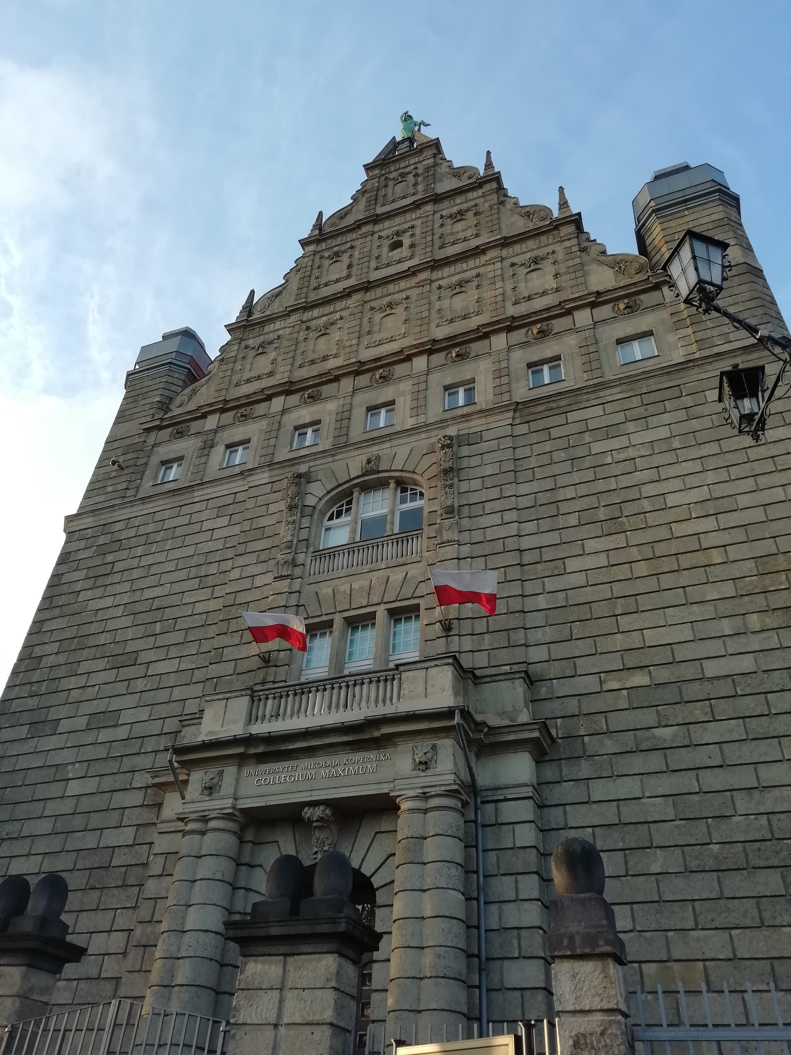 La facciata in mattoni di un edificio storico vista dal basso, con due bandiere polacche al balcone.