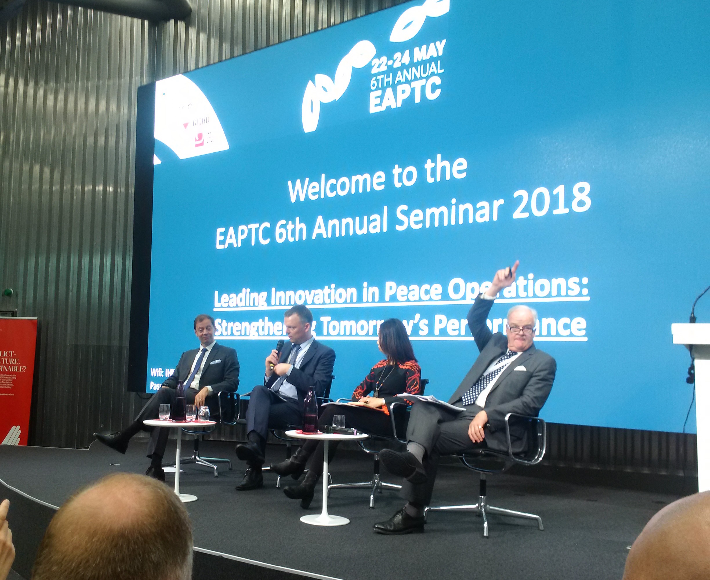 Quattro relatori seduti sul palco davanti alla proiezione Welcome to the EAPTC 6th Annual Seminar 2018, Leading Innovation in Peace Operations.