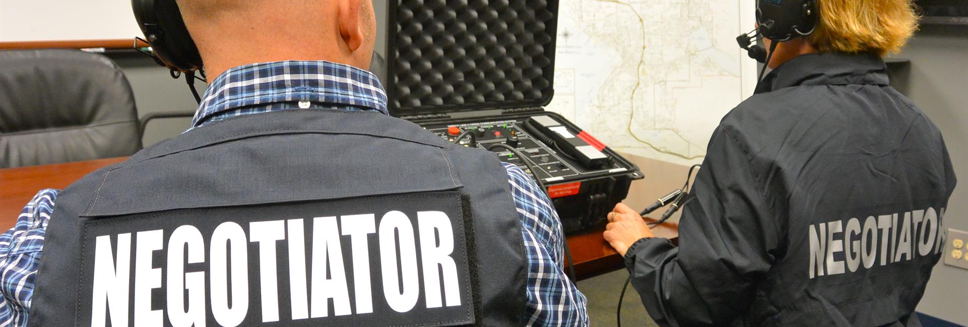 Due persone riprese di spalle con la scritta NEGOTIATOR sul giubbotto e le cuffie in testa, davanti a una valigia con l'apparato radio.