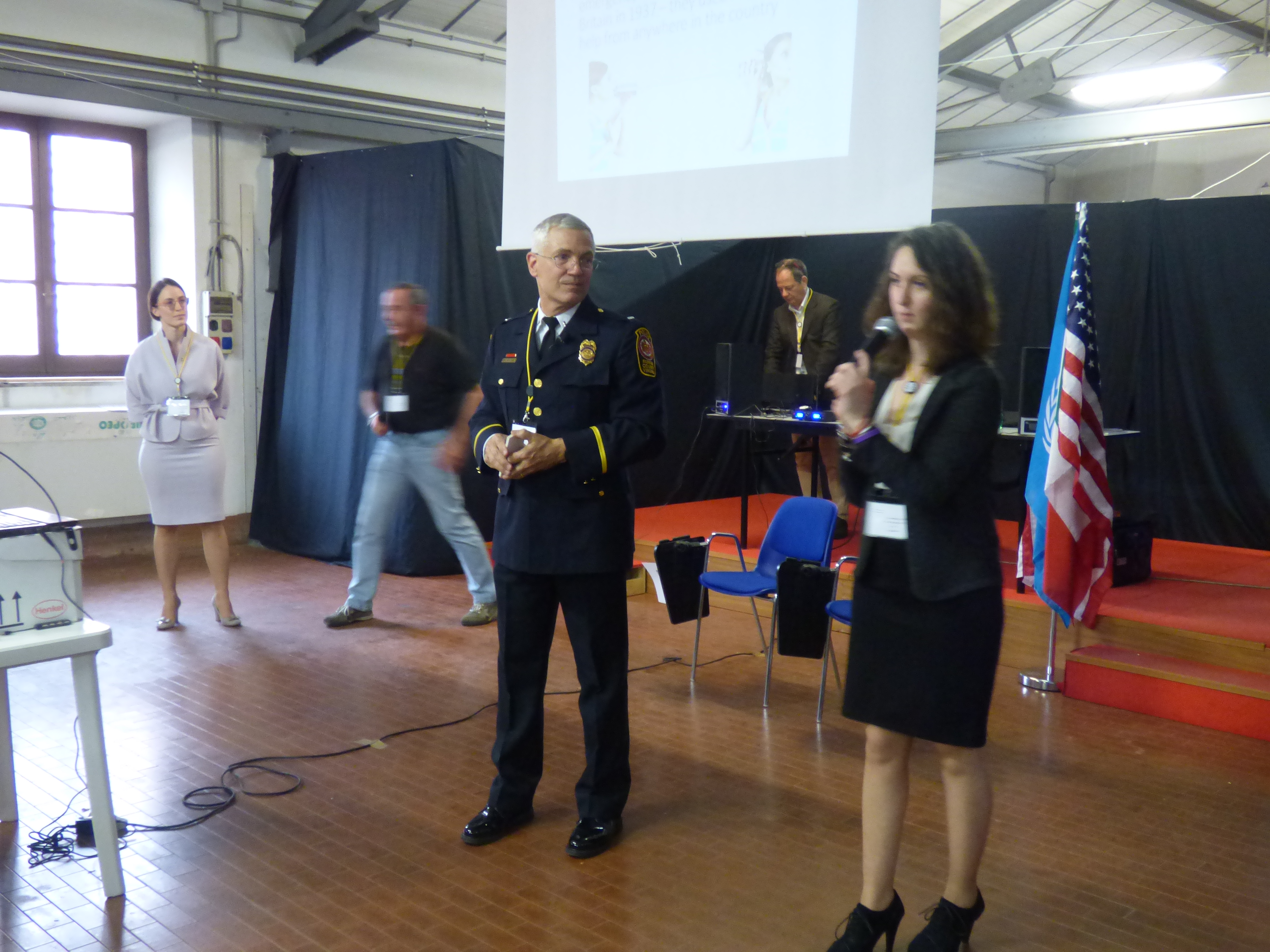 Un ufficiale di polizia statunitense in uniforme accanto a una relatrice che parla al microfono.