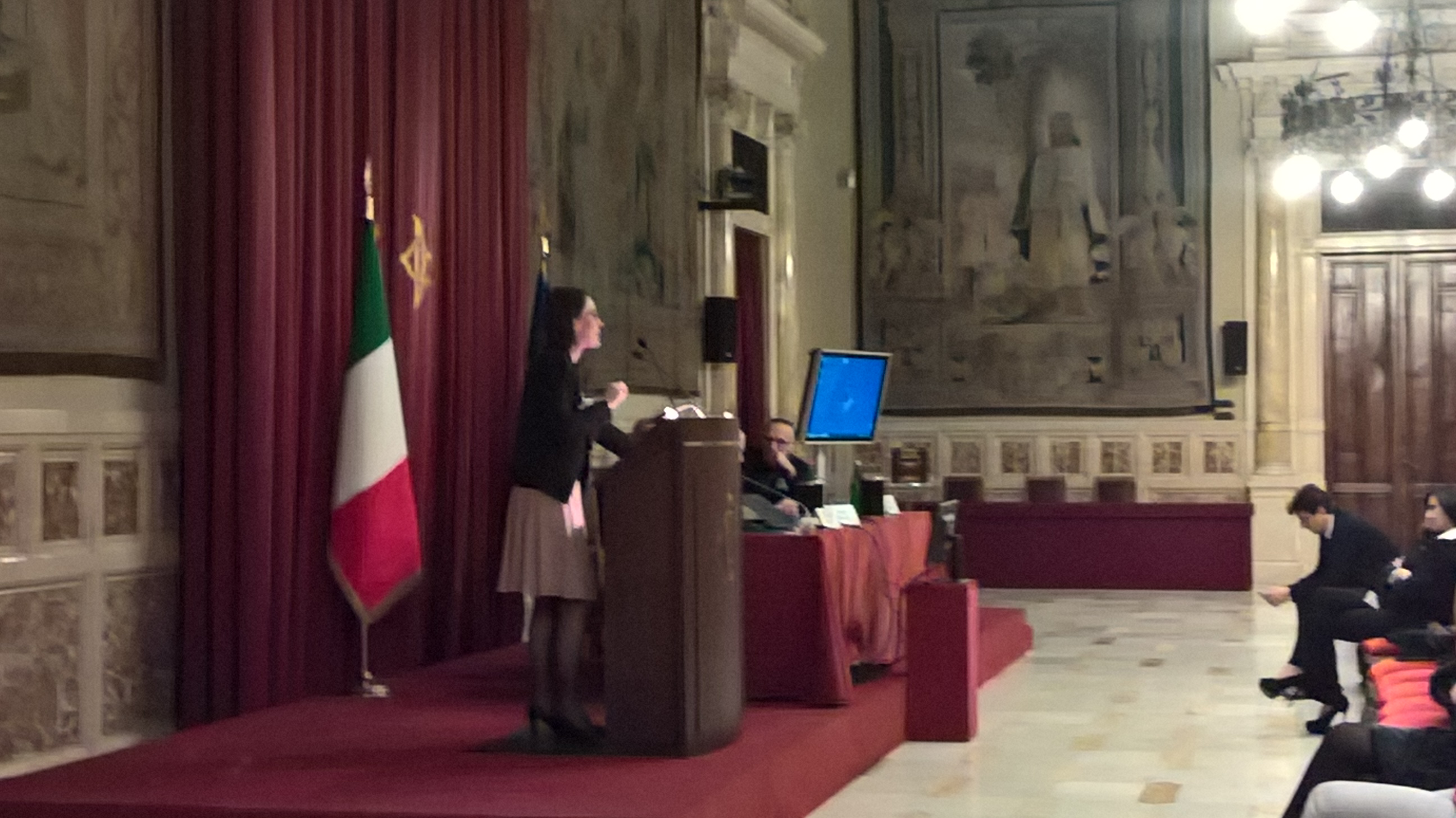 Una relatrice al podio accanto alla bandiera italiana, in una sala parlamentare con i drappi rossi.