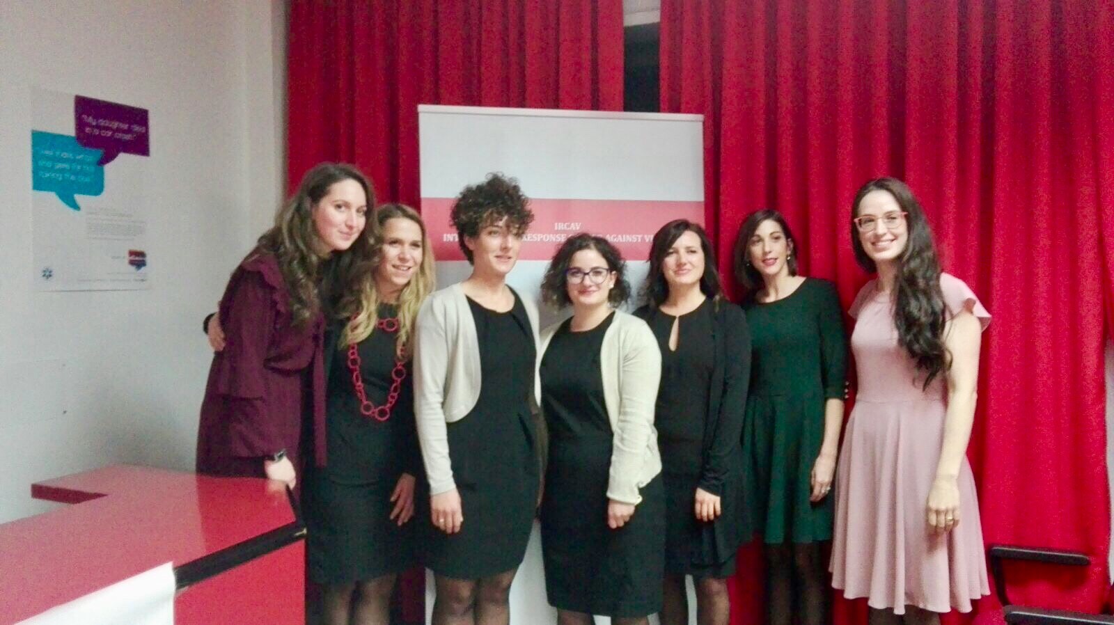 Sette relatrici in posa davanti al pannello della campagna, in una sala dalle tende rosse.