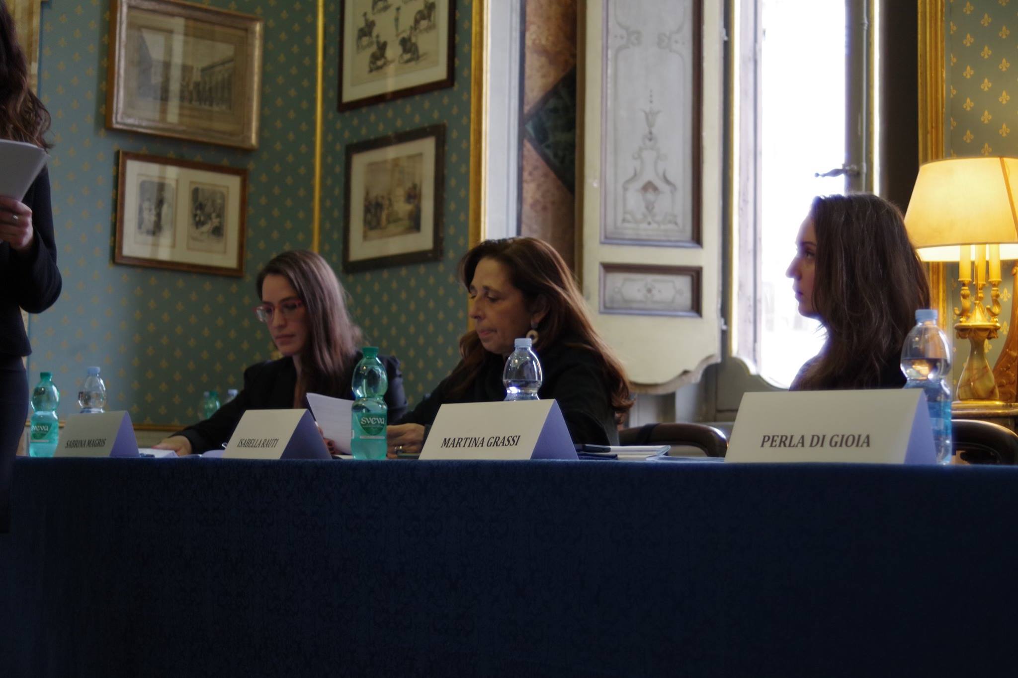 Tre relatrici sedute al tavolo con i cartellini dei nomi, in una sala con i quadri alle pareti.
