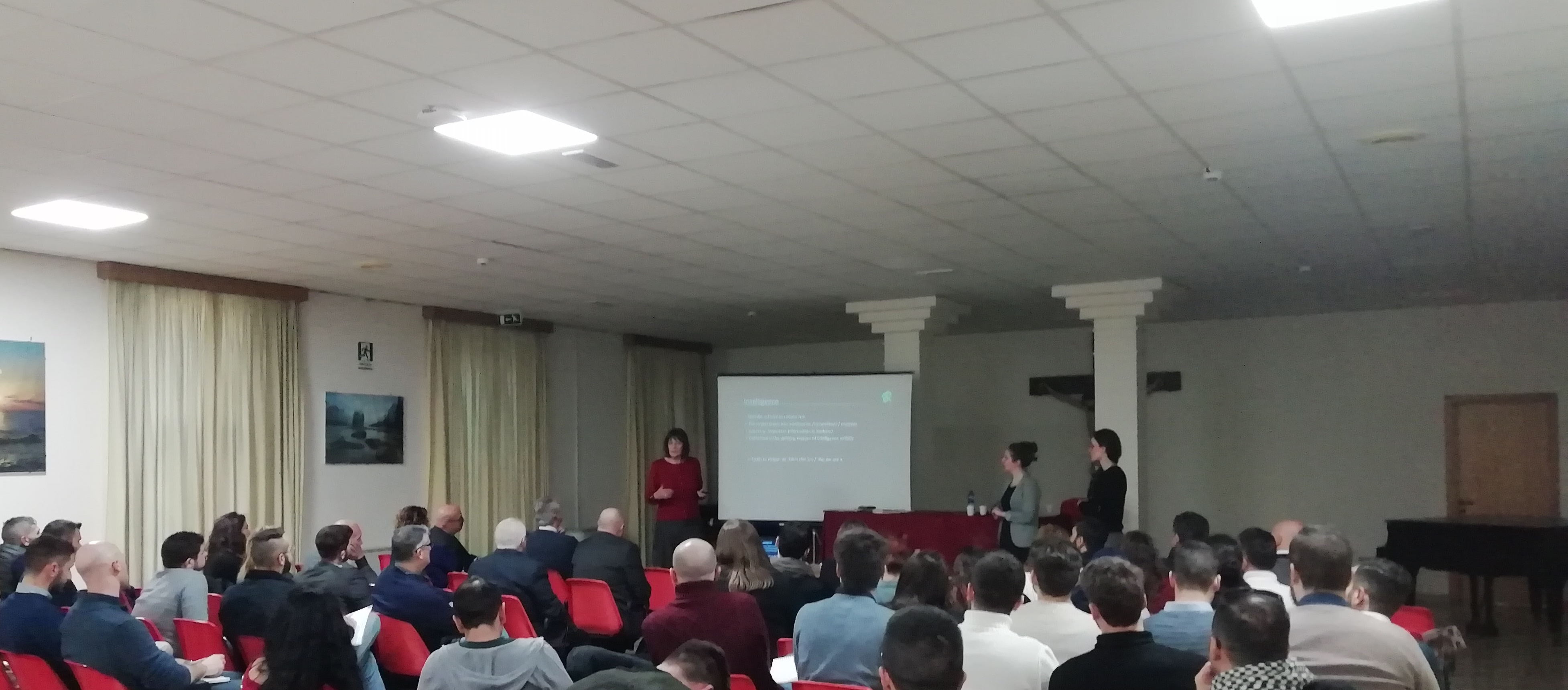 La sala piena vista da dietro, con tre relatori davanti allo schermo.