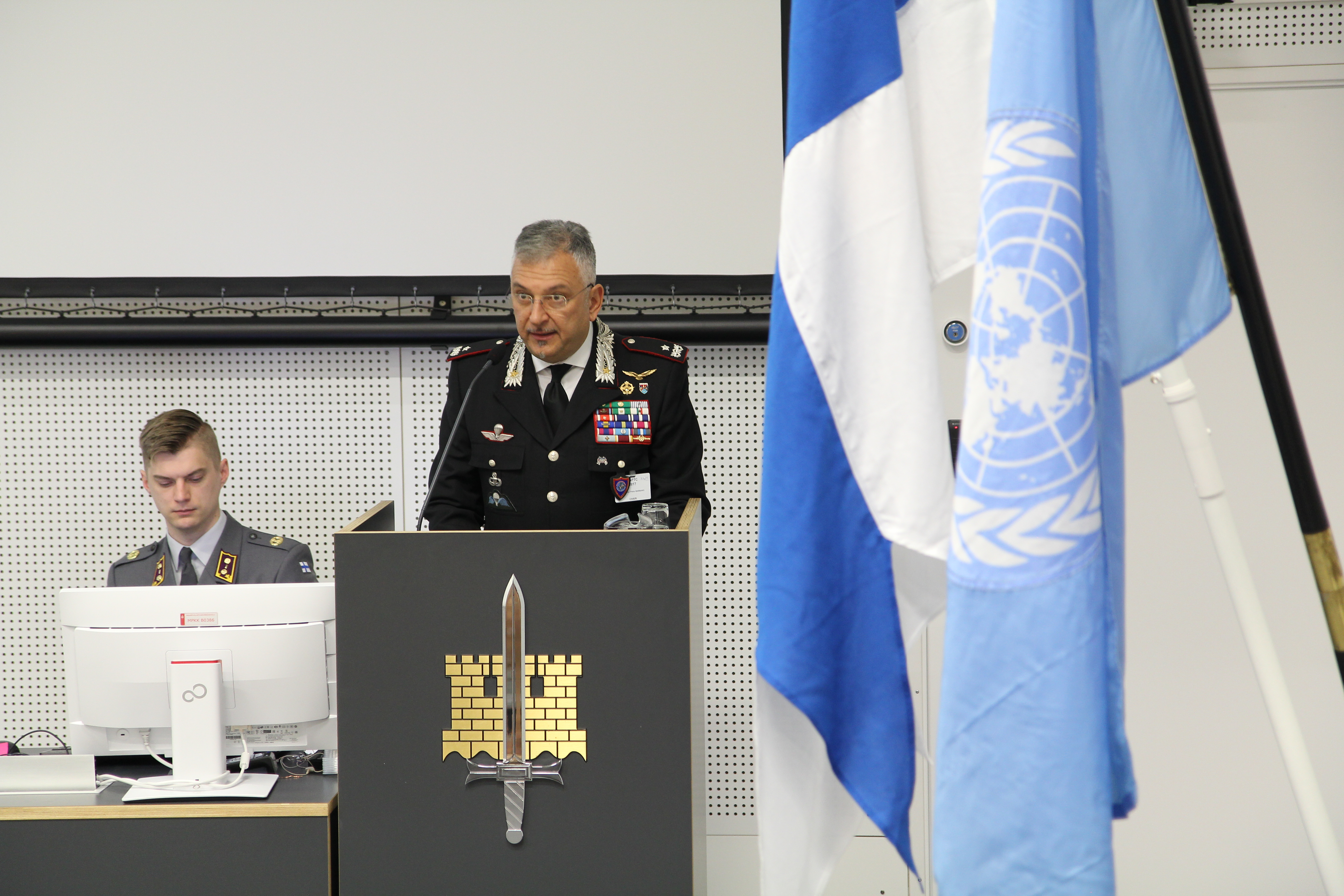 Un ufficiale dei carabinieri parla dal podio, accanto alle bandiere finlandese e delle Nazioni Unite.