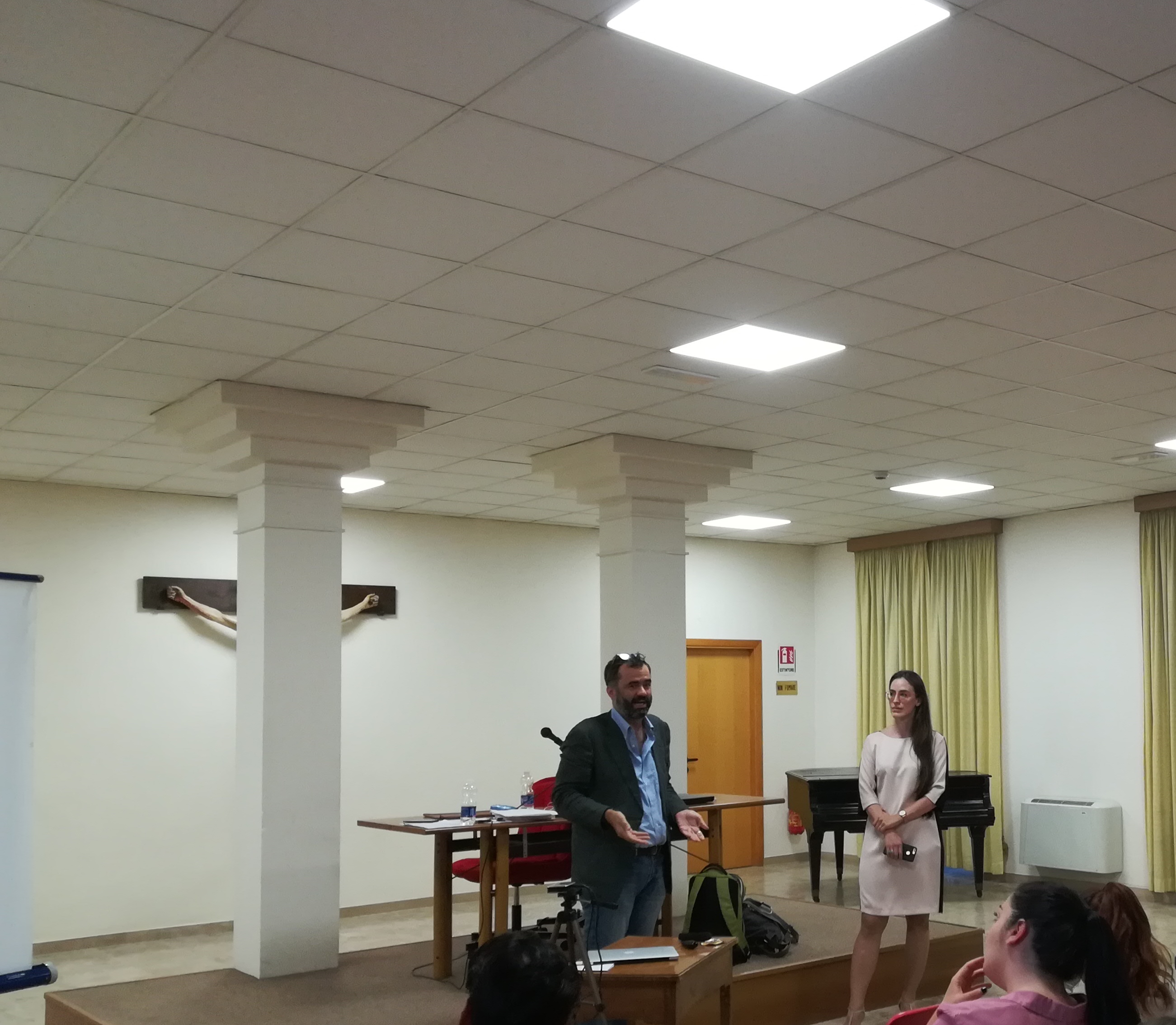 Un relatore e una relatrice in piedi davanti al pubblico, in una sala con le colonne e il pianoforte.