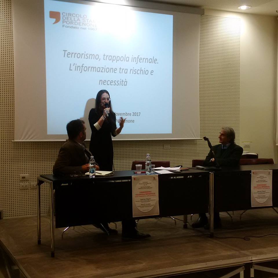 Una relatrice al microfono davanti alla proiezione Terrorismo, trappola infernale. L'informazione tra rischio e necessità, con due persone al tavolo.