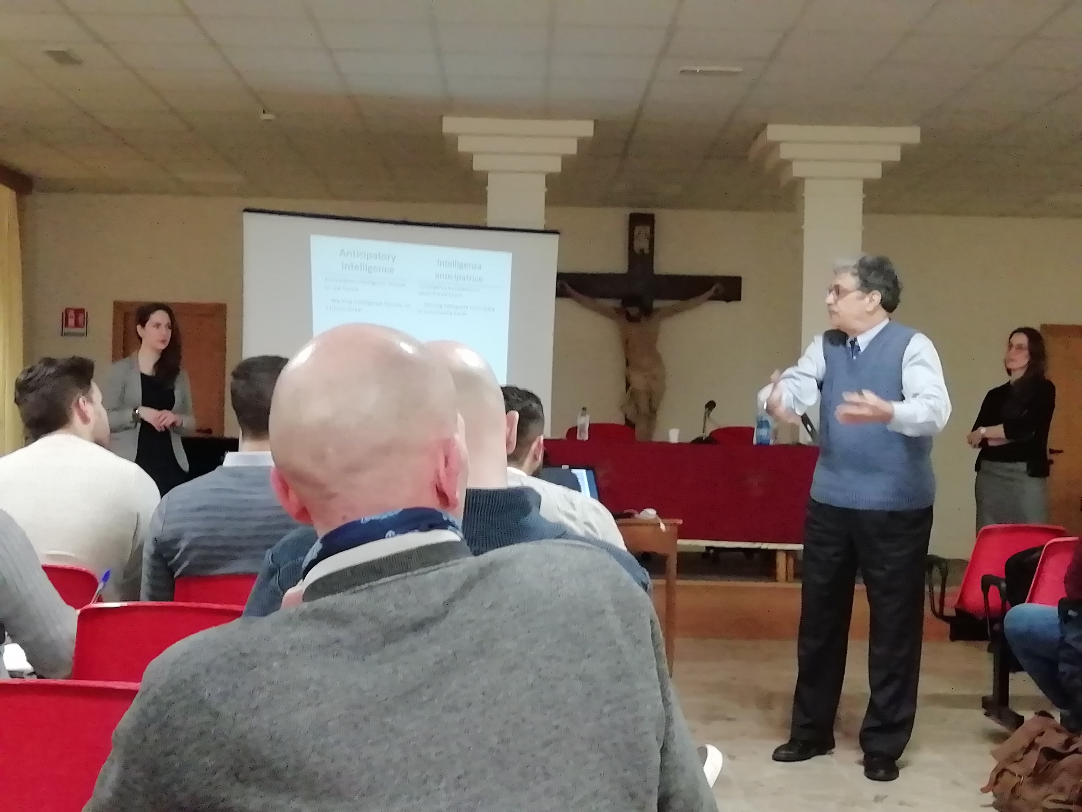 Un relatore parla in piedi fra il pubblico, con due relatrici ai lati della sala.