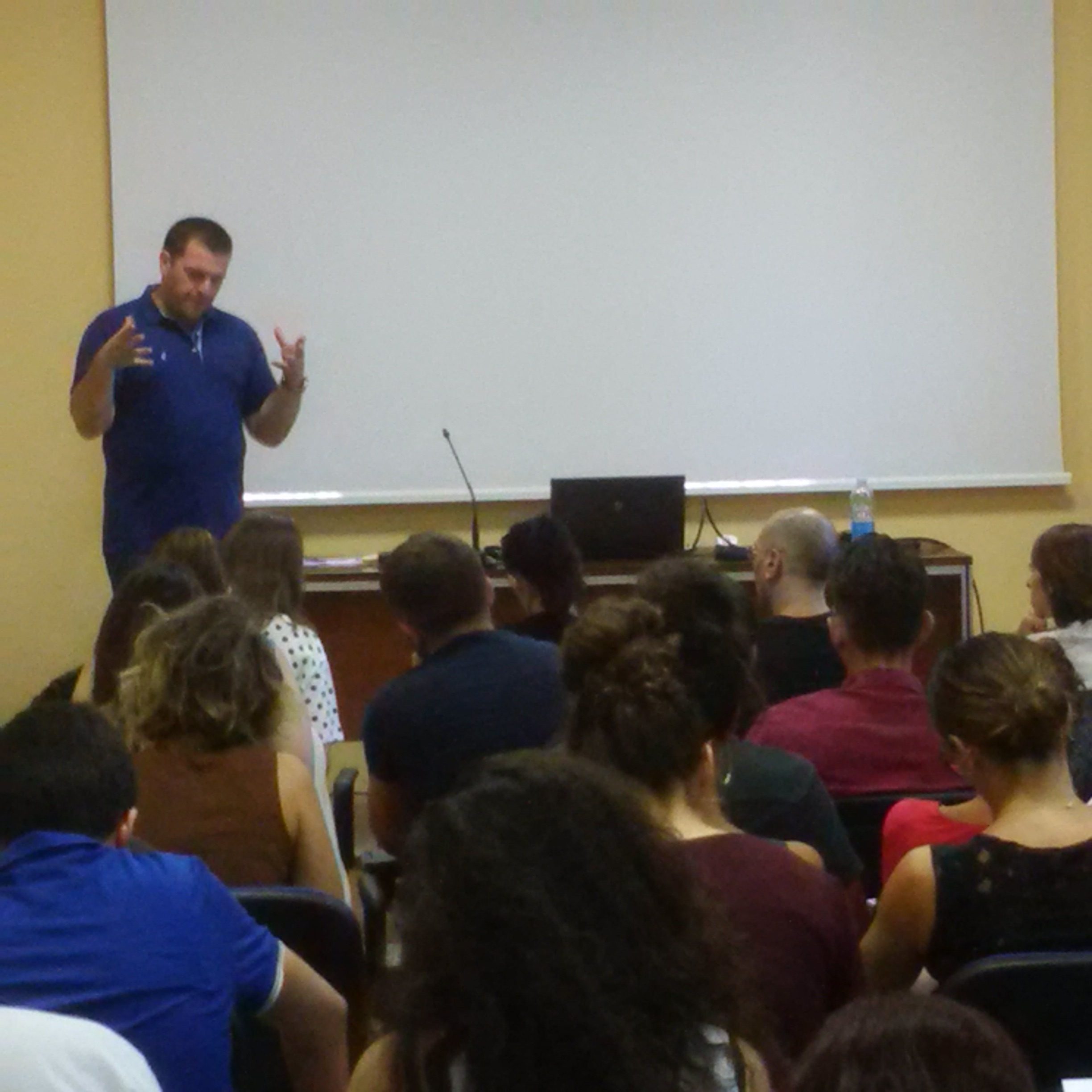 Un relatore in polo blu parla davanti al pubblico seduto, ripreso da dietro le sedie.