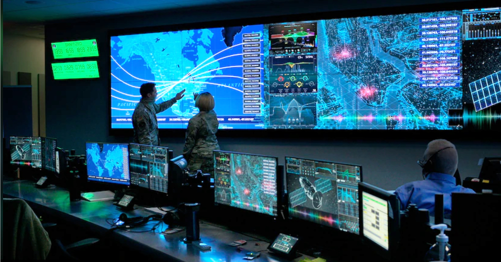 Una sala di controllo: due persone davanti a un grande schermo con mappe e grafici, attorno postazioni con molti monitor.