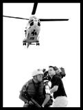 Fotografia in bianco e nero: un elicottero di soccorso in volo, sotto due soccorritori sostengono una persona ferita.