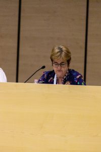 Una relatrice al microfono dietro il banco di legno.