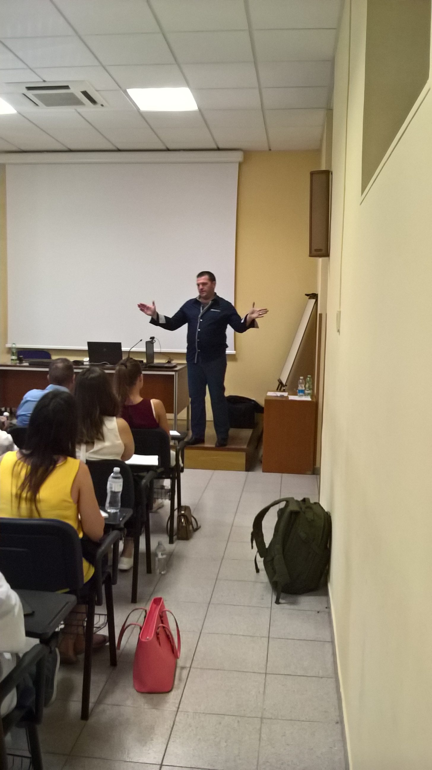 Un relatore con le braccia aperte parla su una pedana davanti al pubblico seduto.