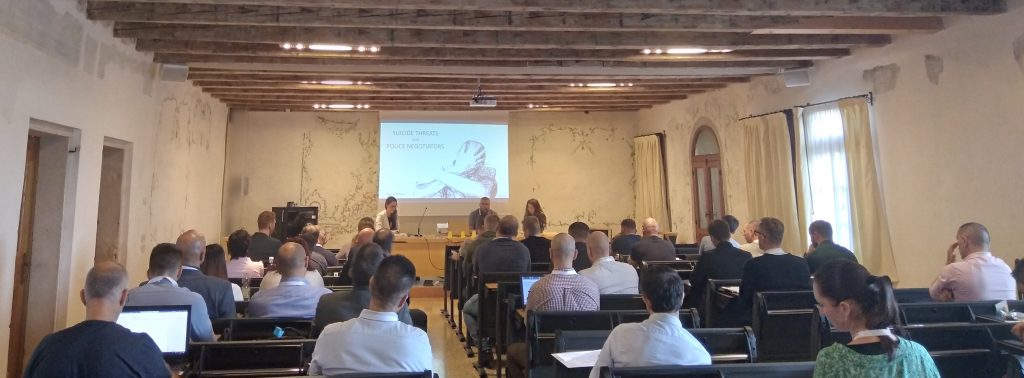 Una sala con il soffitto a travi di legno: i partecipanti seduti e due relatori accanto allo schermo.
