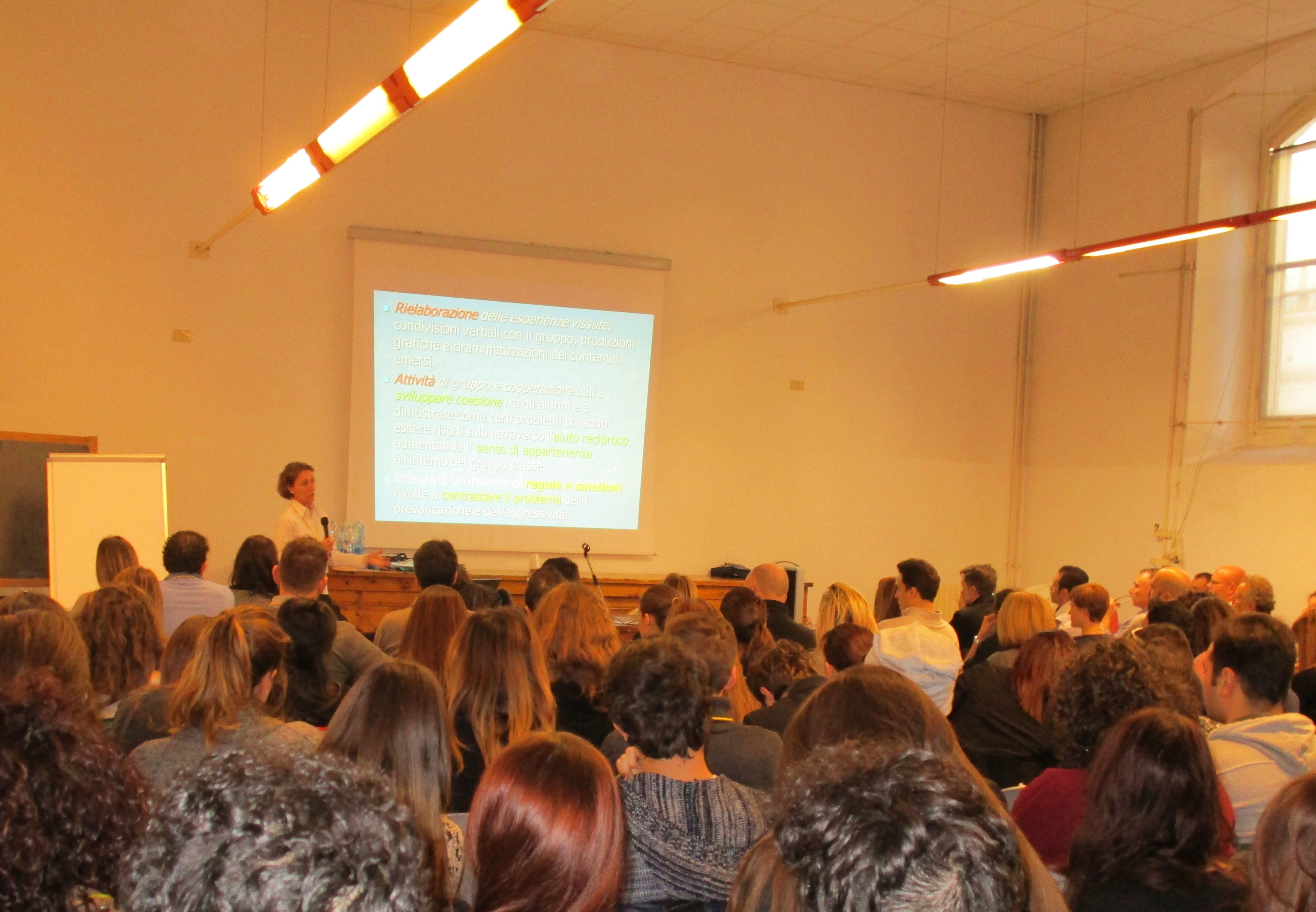 Un'aula piena vista da dietro; in fondo una relatrice accanto alla proiezione delle diapositive.