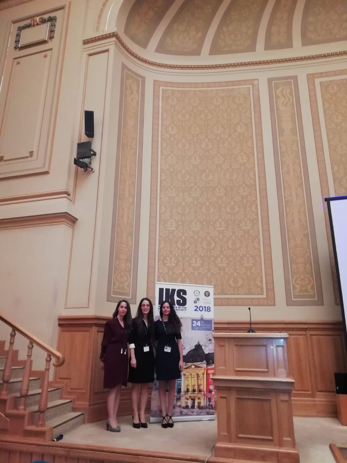 Tre partecipanti in posa davanti al banner IKS 2018, in un'aula magna decorata.