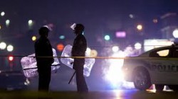Di notte, due agenti oltre il nastro che delimita una scena del crimine, illuminati dai lampeggianti di un'auto di polizia.
