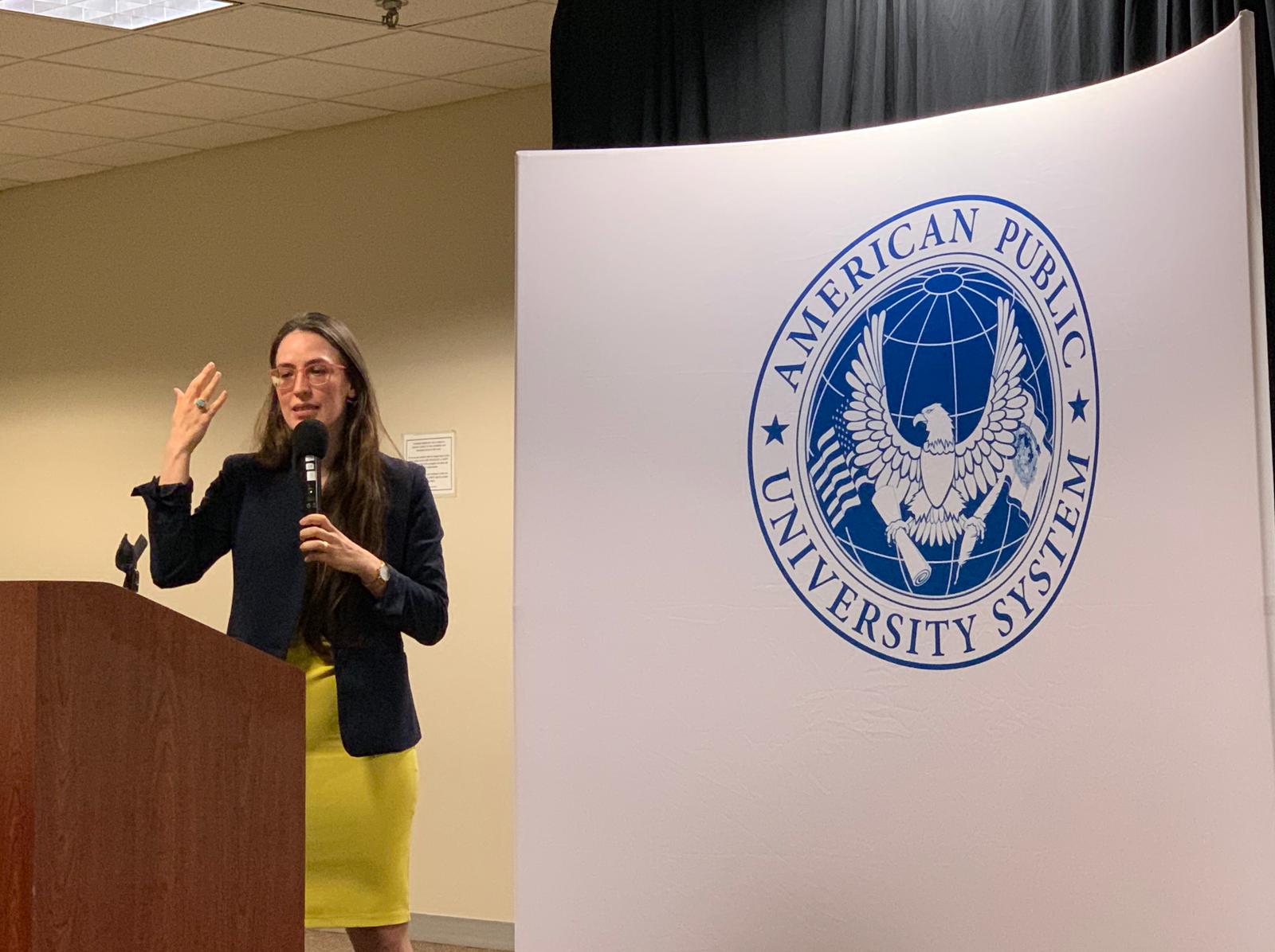 Una relatrice parla al microfono accanto al pannello con il sigillo dell'American Public University System.
