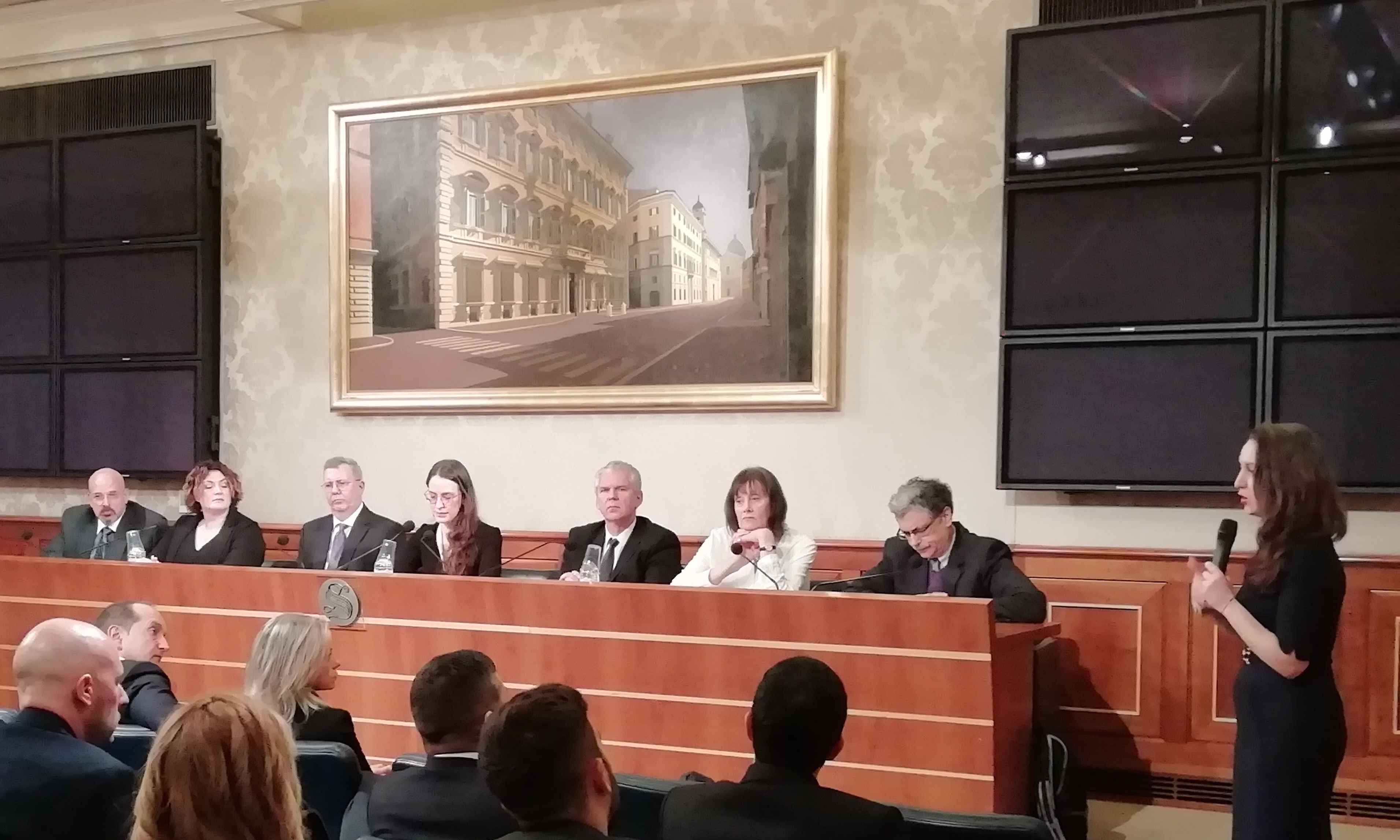 La stessa sala durante i lavori: una relatrice in piedi parla ai sette al tavolo e al pubblico.
