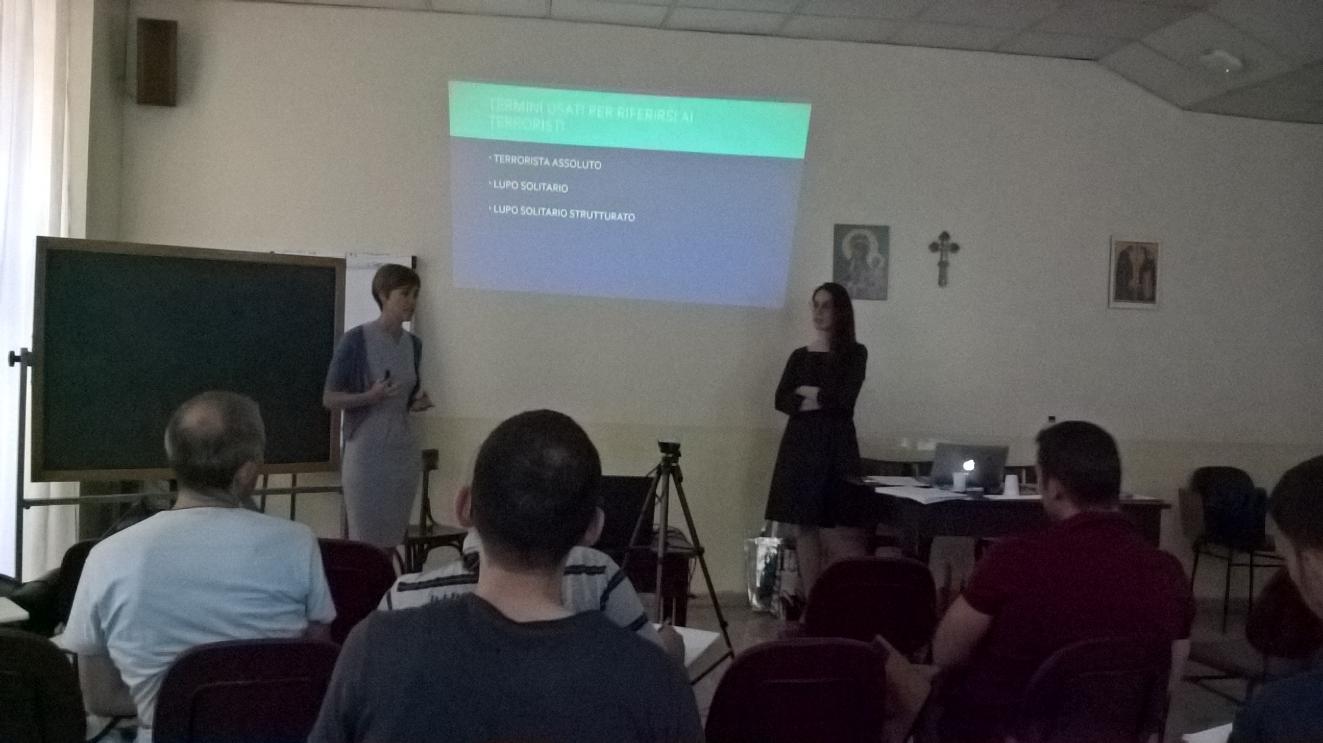 Due relatrici accanto alla proiezione di una diapositiva, in un'aula con il pubblico seduto.