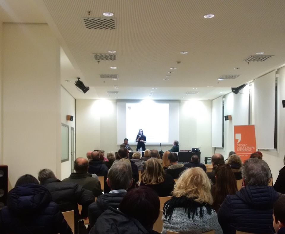 La sala piena vista da dietro, con la relatrice al tavolo sotto lo schermo.